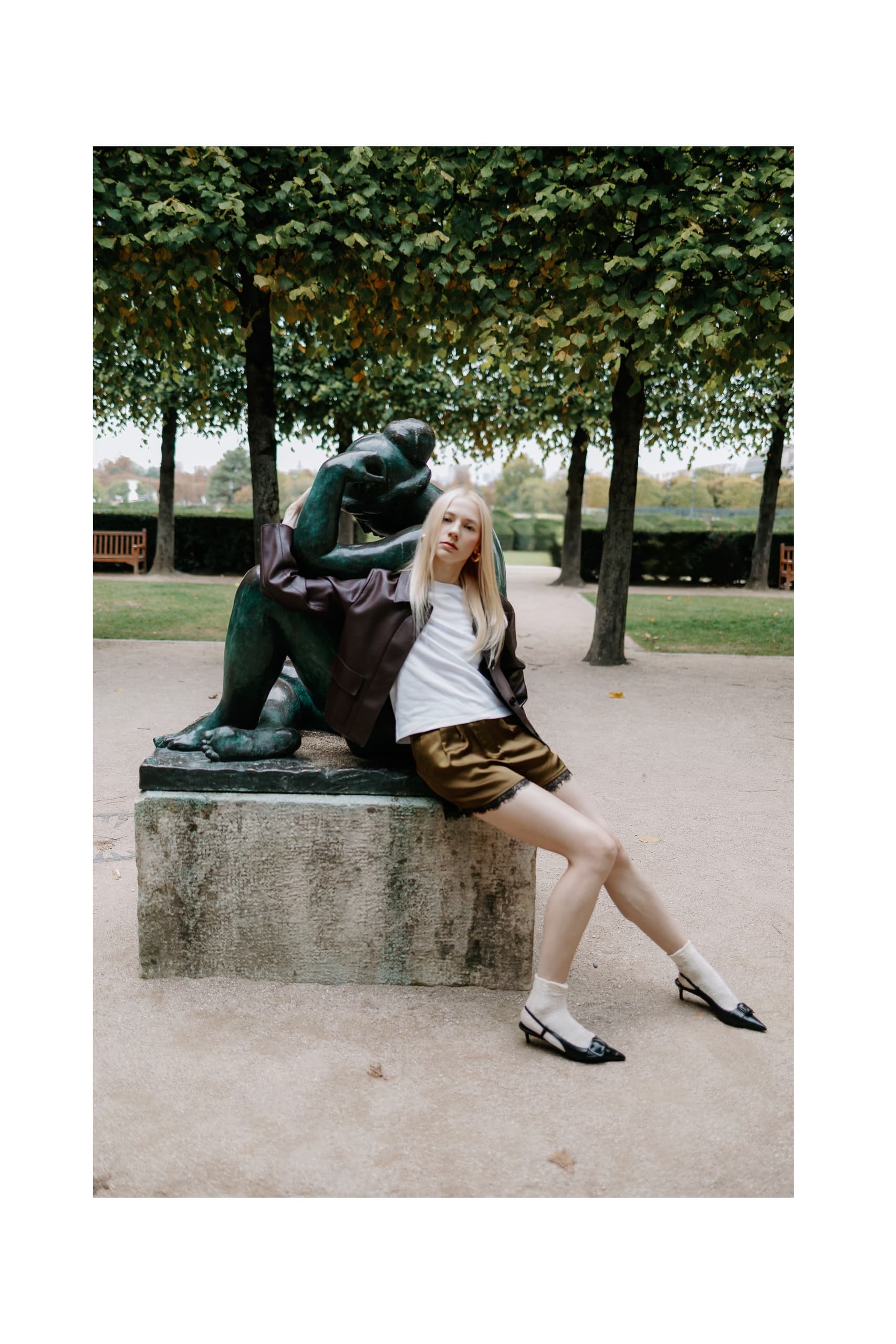 Fashionable woman poses by garden statue in stylish outfit.