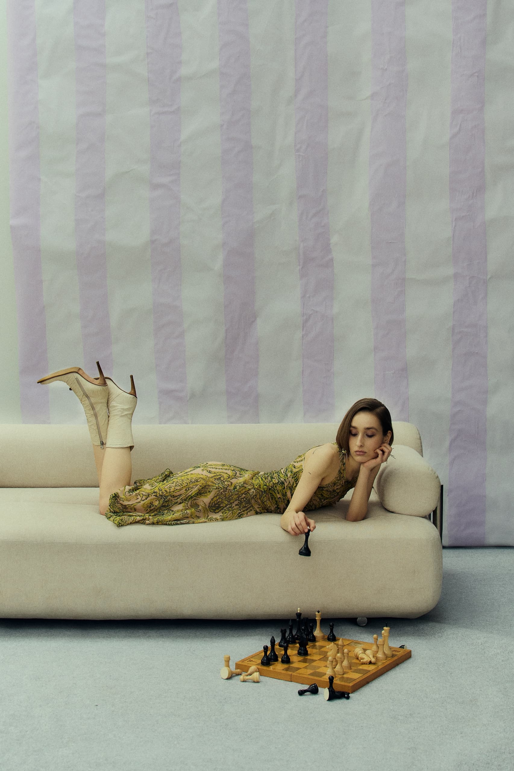 Woman playing chess while lying on a couch in a patterned dress.