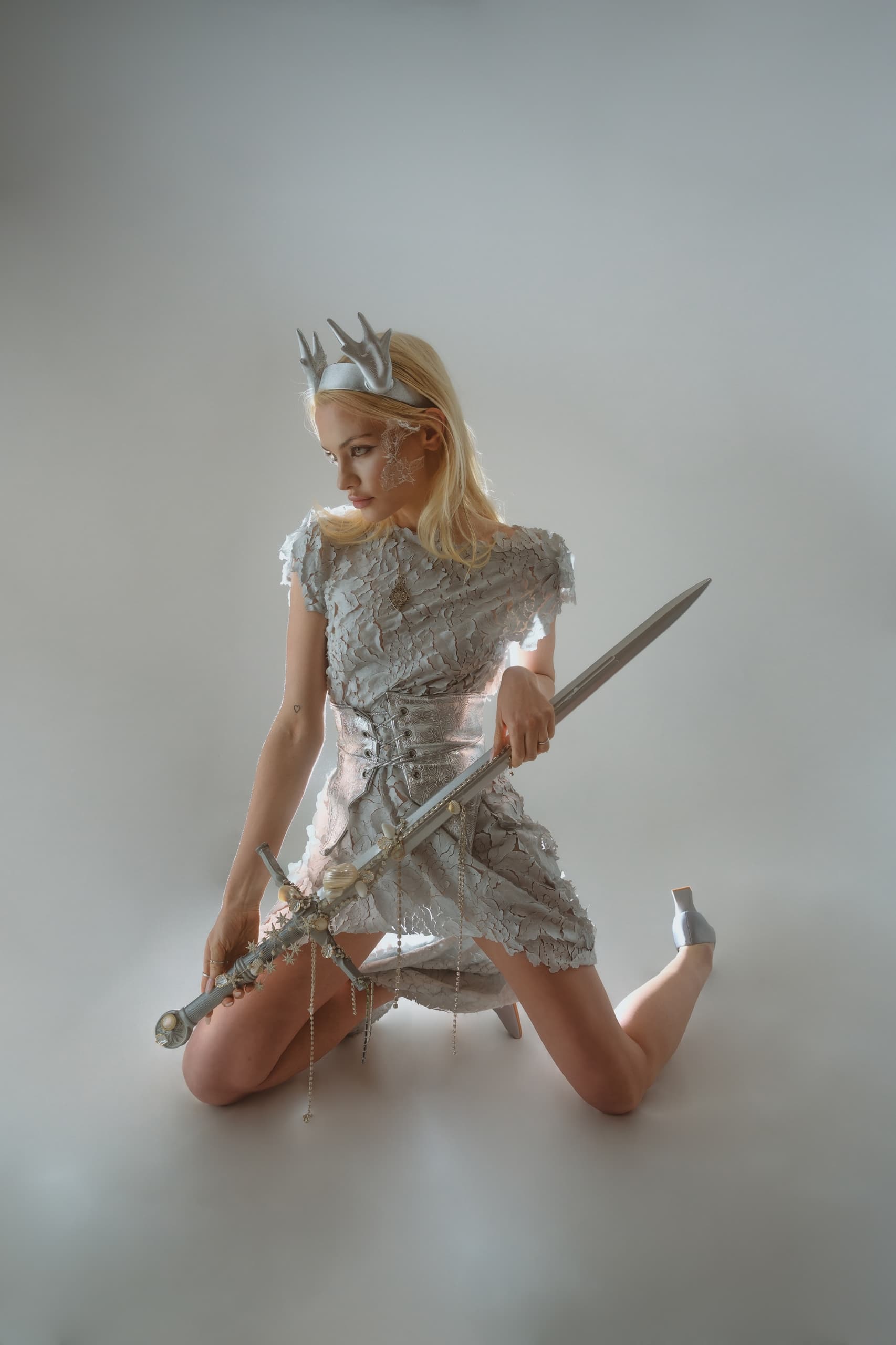 Fantasy warrior princess with crown and sword in silver attire kneeling on the ground.