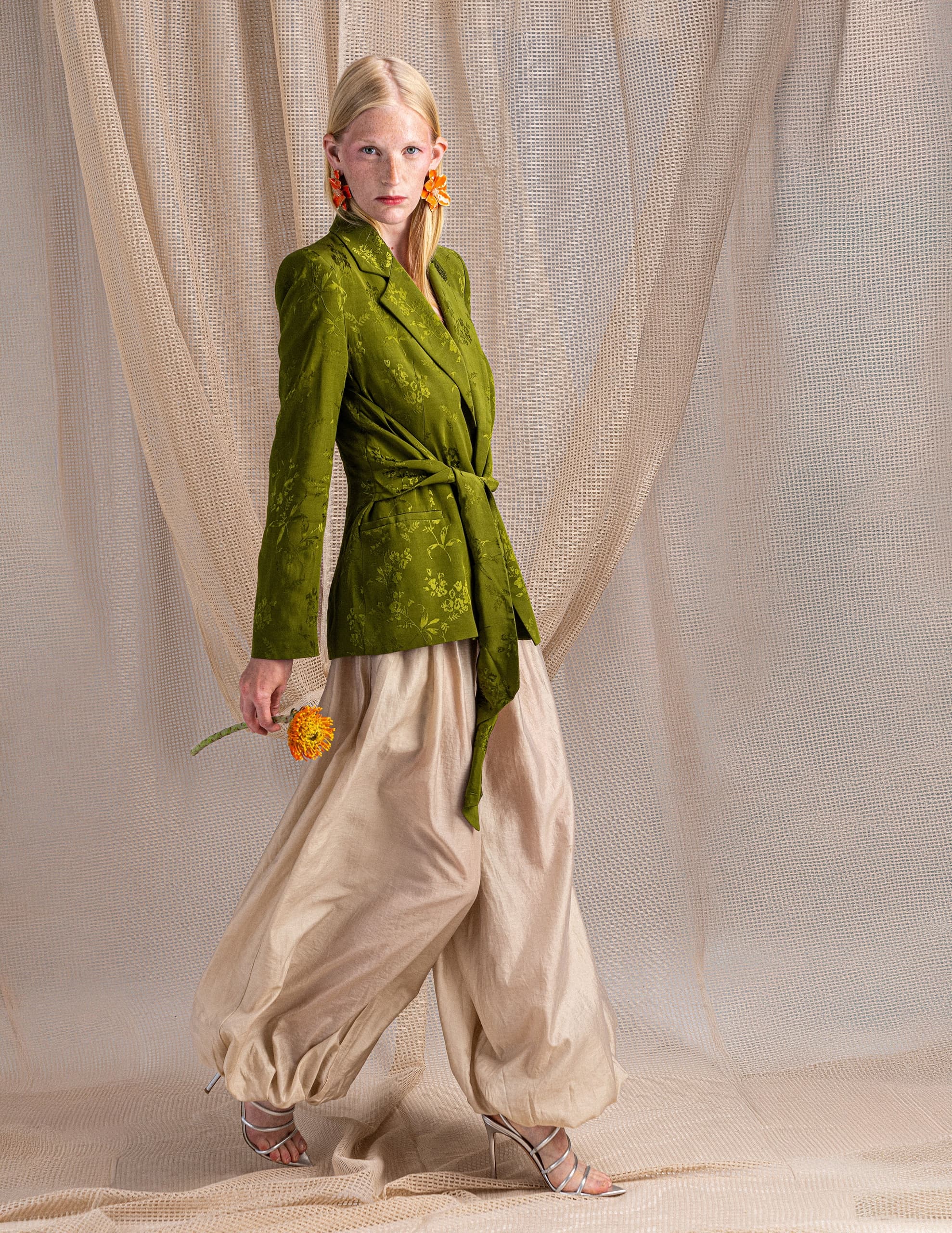 Model in green blazer and beige pants holds a flower in a stylish fashion photoshoot.