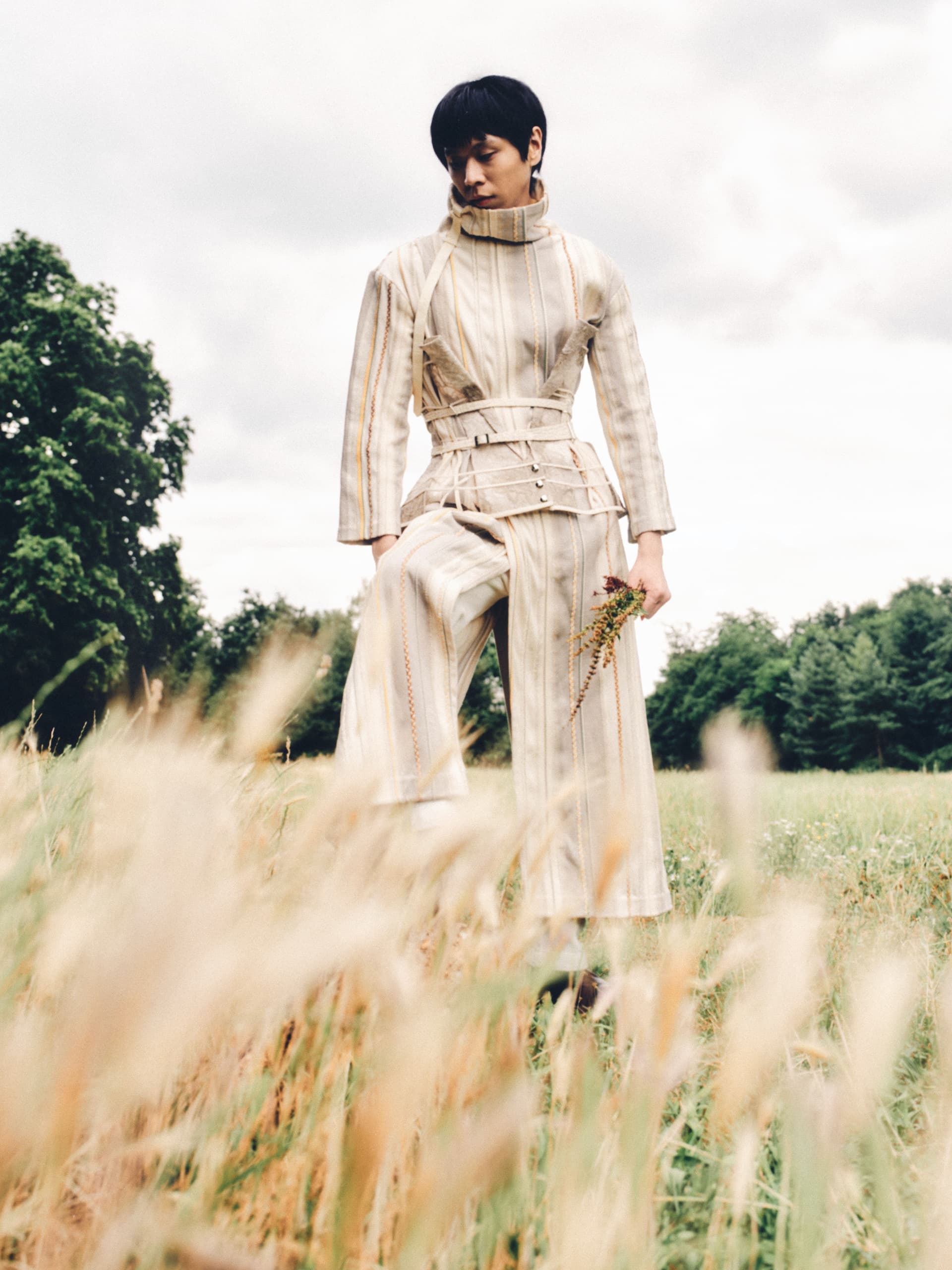 Person in a stylish outfit holding flowers in a scenic grassy field.