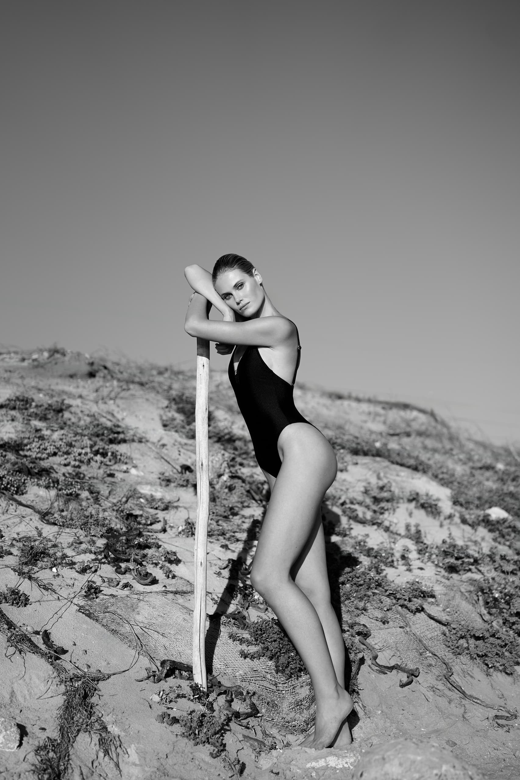 Woman in a swimsuit leaning on a stick, posing on a sandy beach.