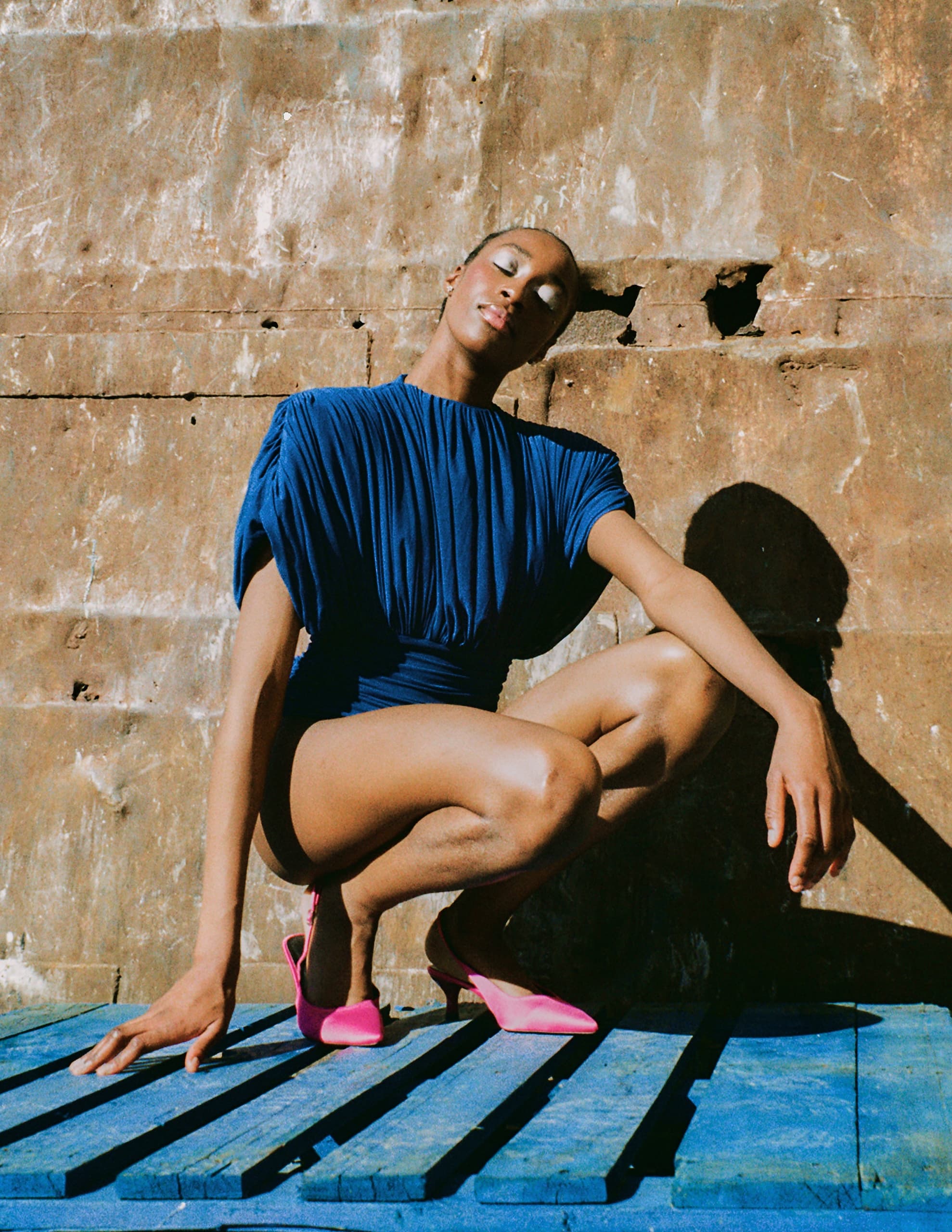 Person in blue outfit poses stylishly on a wooden platform with pink heels.