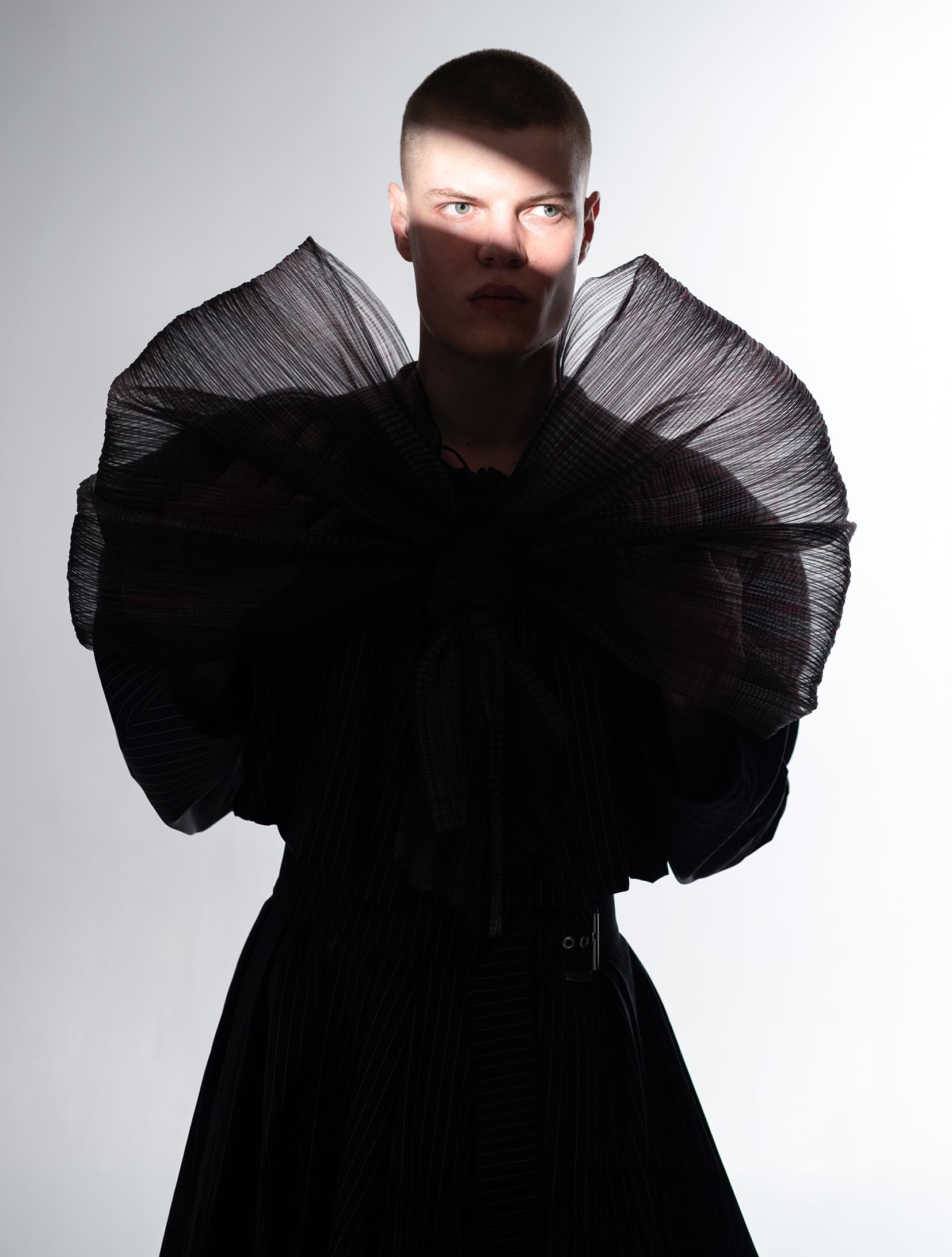 Person in dramatic black attire with a bold, structured collar in moody lighting.