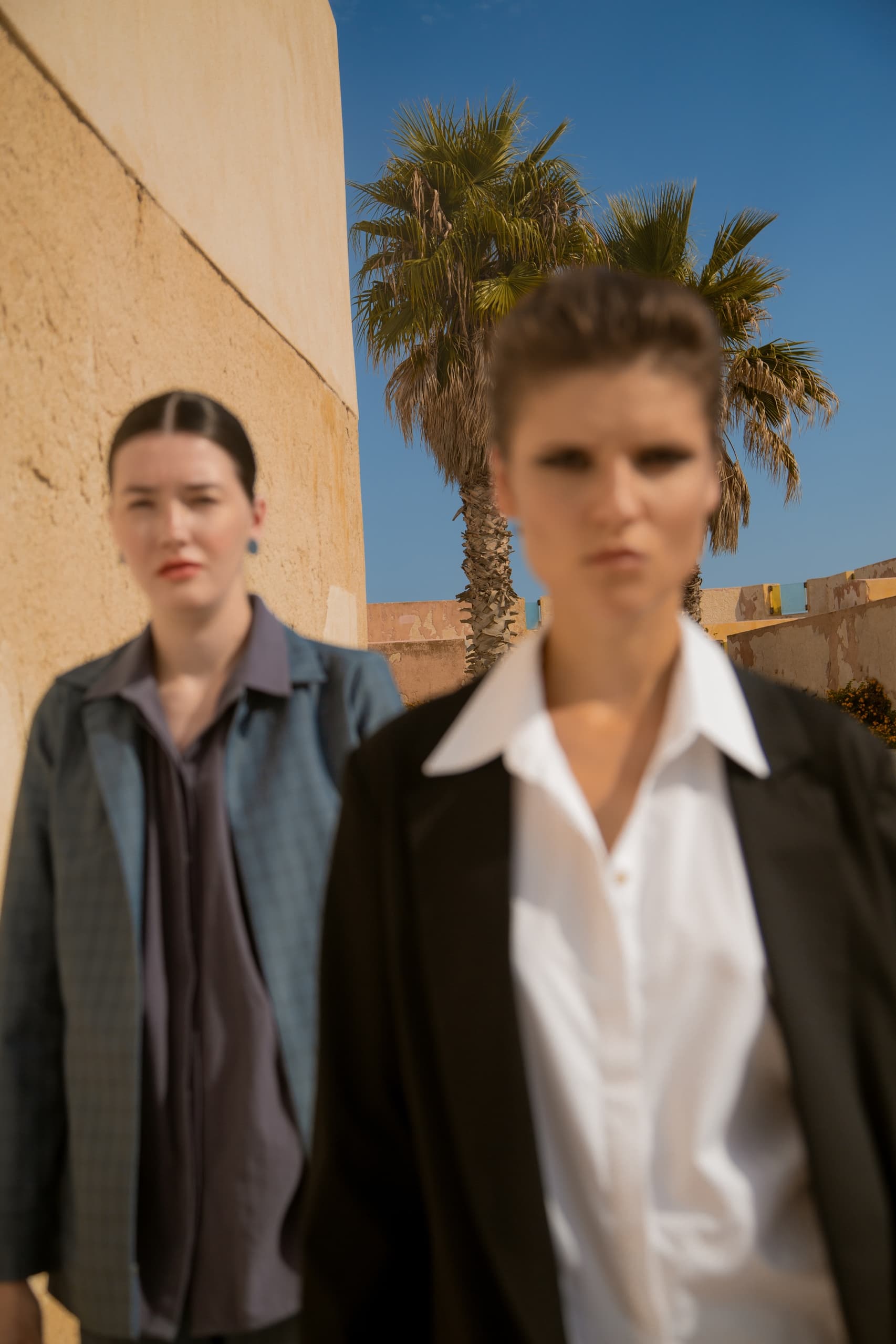 Two women in business attire with a palm tree and blue sky background.