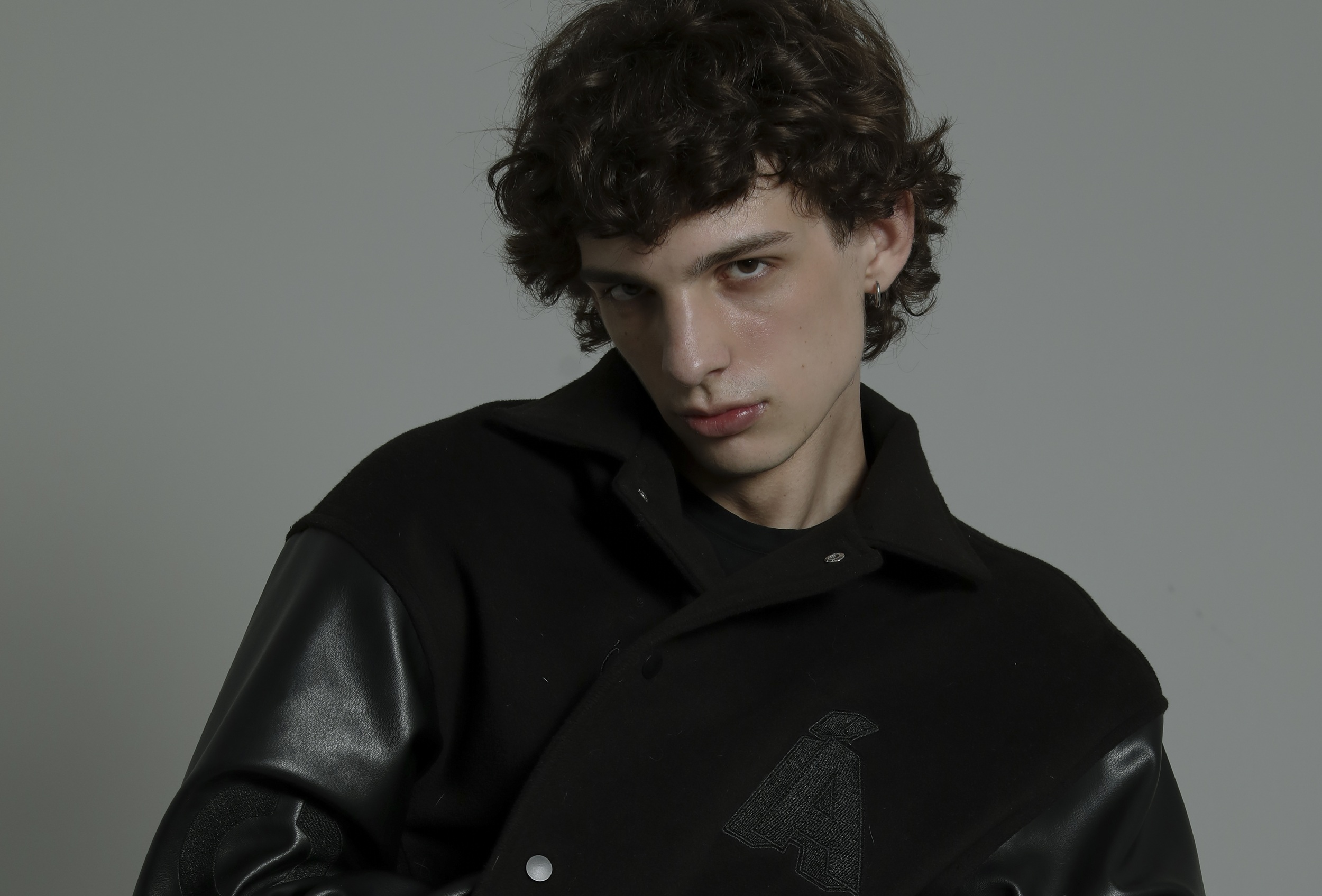 Curly-haired person in stylish black jacket posing confidently against gray background.