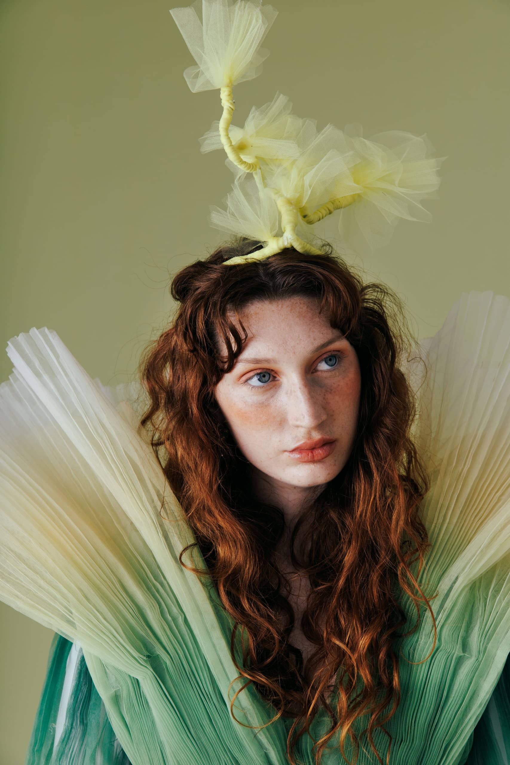 Model in green tulle dress with creative headpiece, red curly hair, green background.