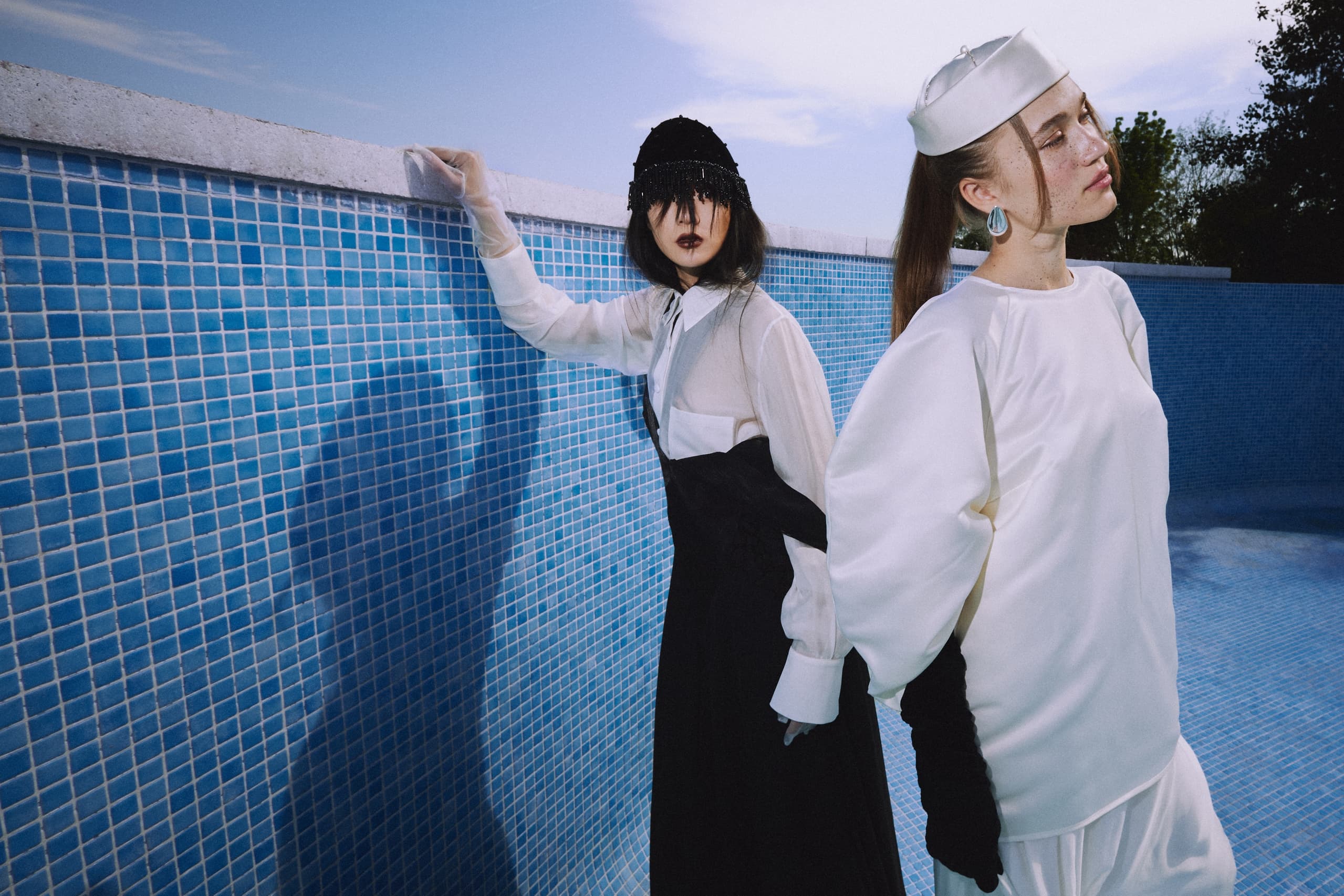 Two models in avant-garde fashion pose by a blue tiled wall under a clear sky.
