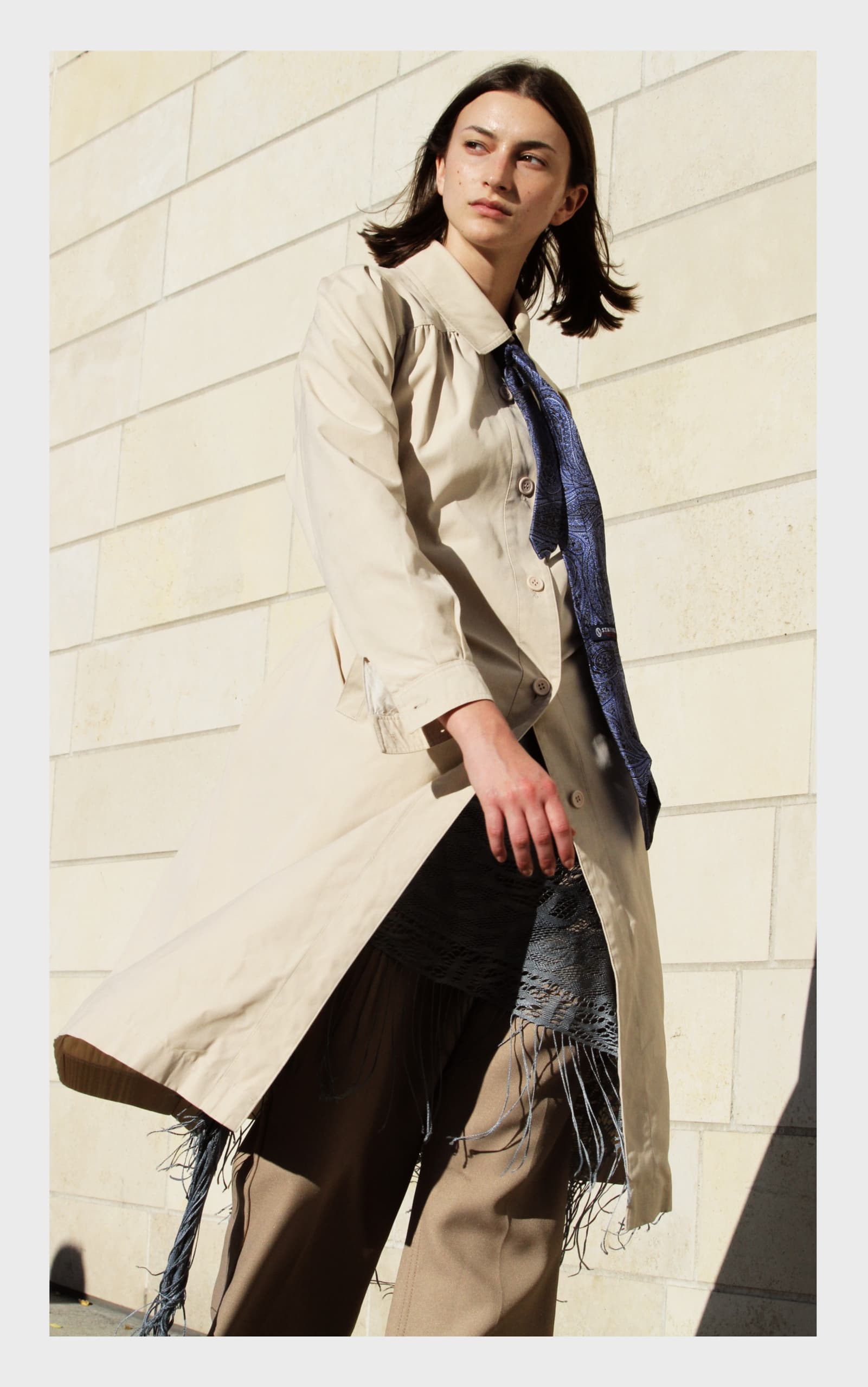 Woman in a beige trench coat and fringed outfit against a light brick wall.