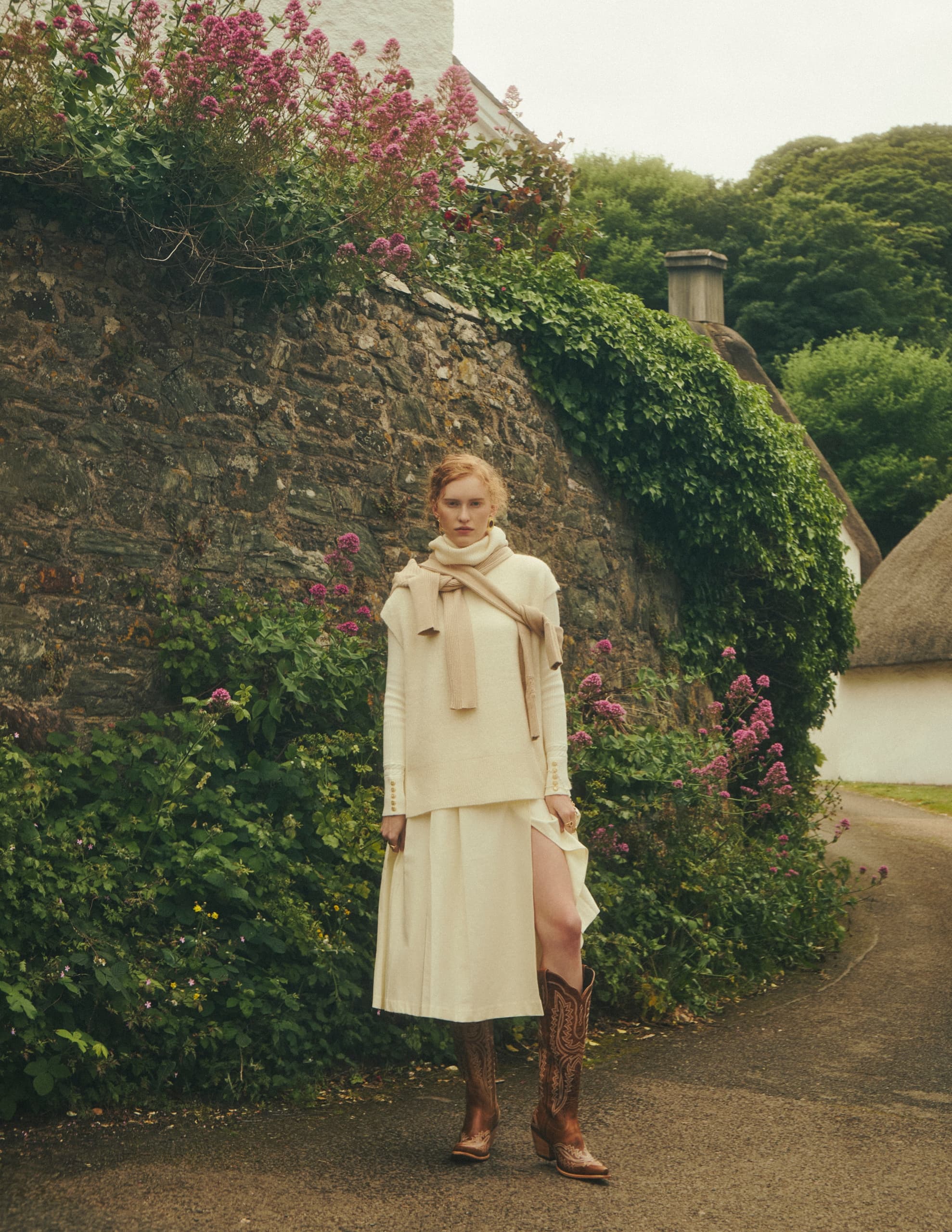 Woman in cream outfit with boots stands by a stone wall adorned with pink flowers.