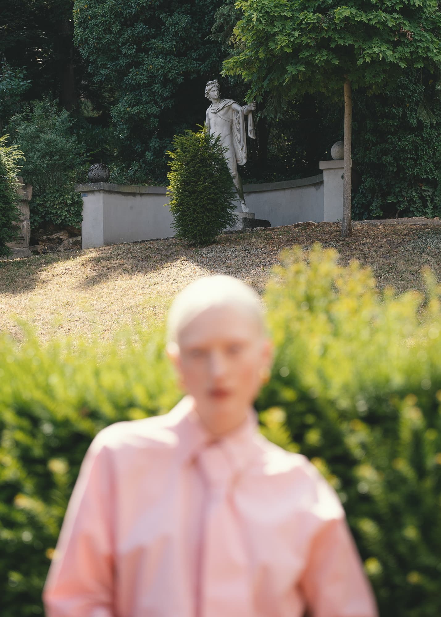 Blurred person in pink before garden statue amidst green foliage.
