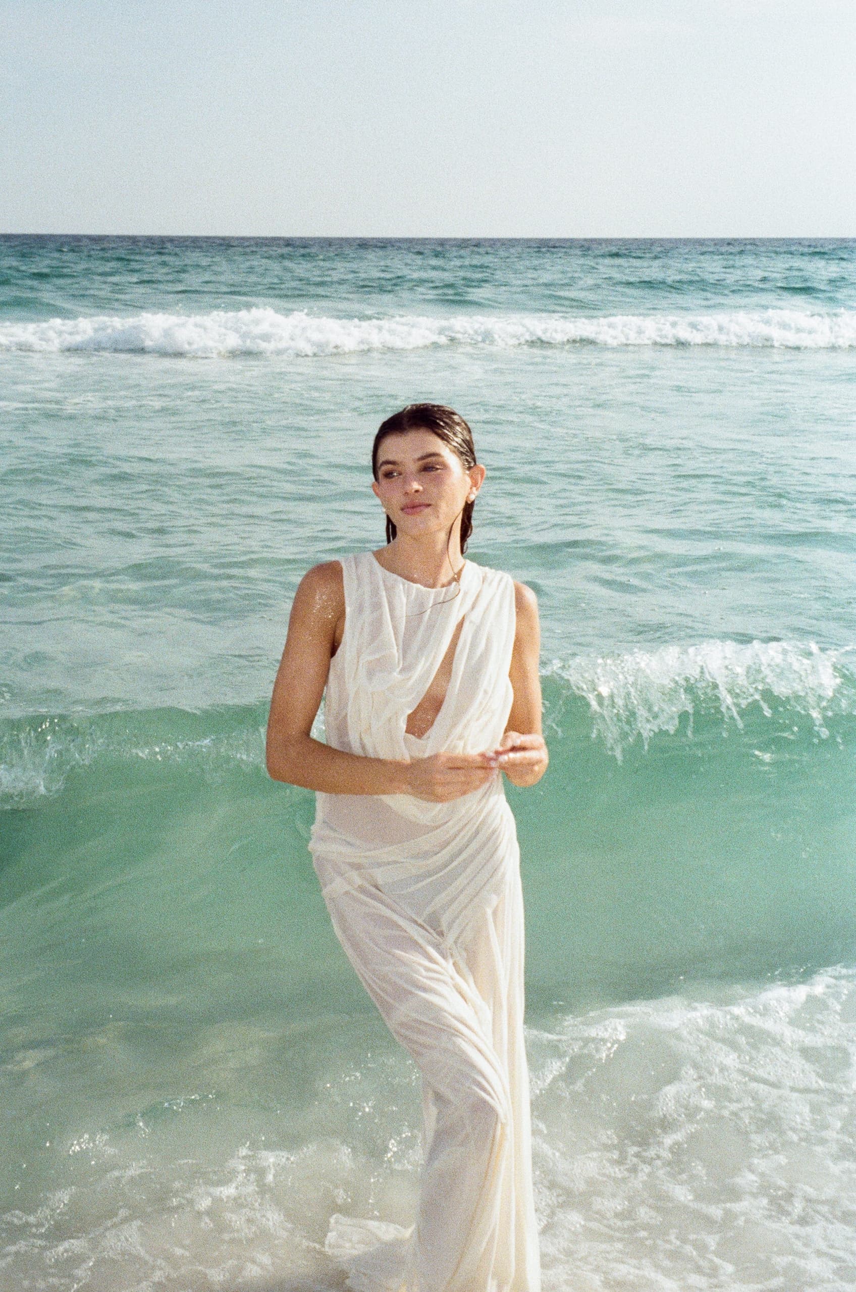 Woman in flowing white dress stands in ocean waves under a bright, clear sky.