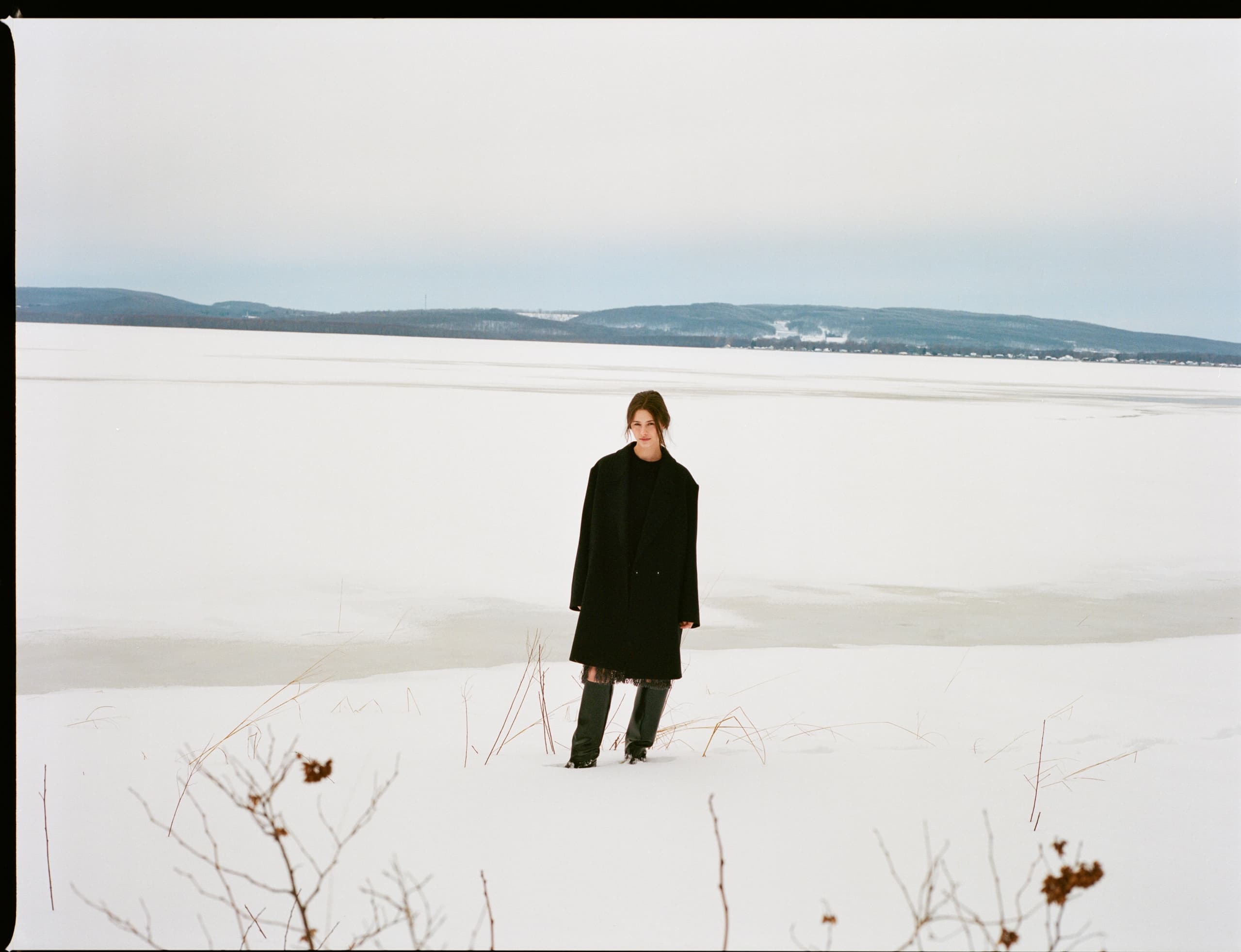 Person in black coat stands on snowy landscape by frozen lake.