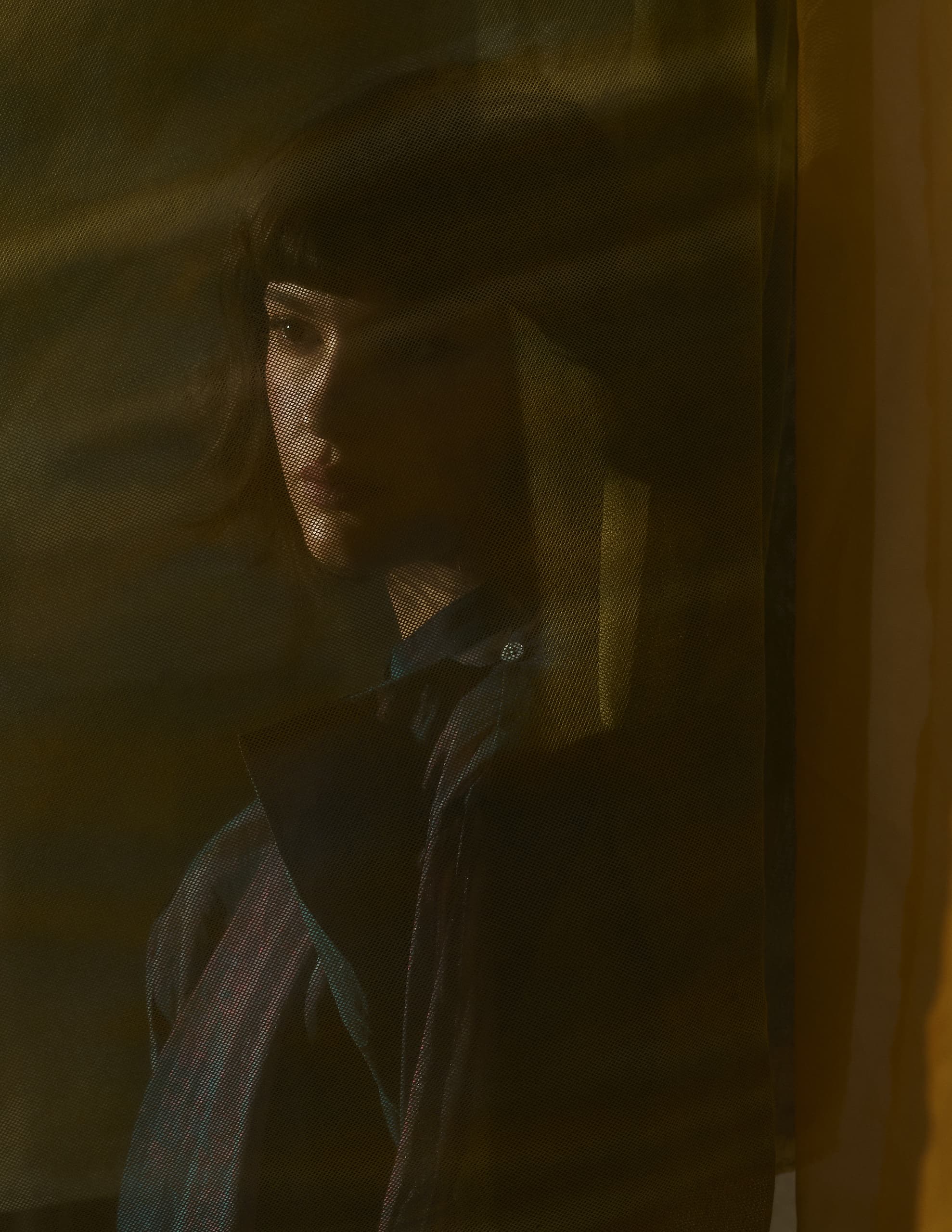 Mysterious figure behind sheer curtain in dimly lit, shadowy setting.