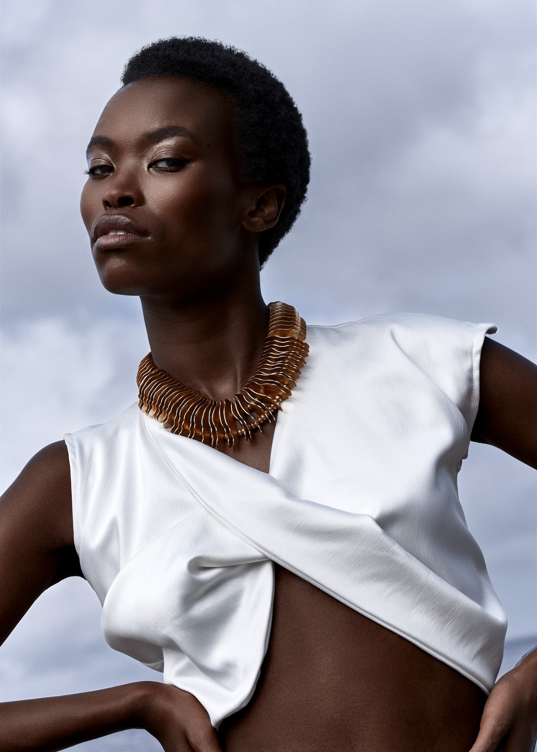 Fashion model in white top and statement necklace against a cloudy sky backdrop.