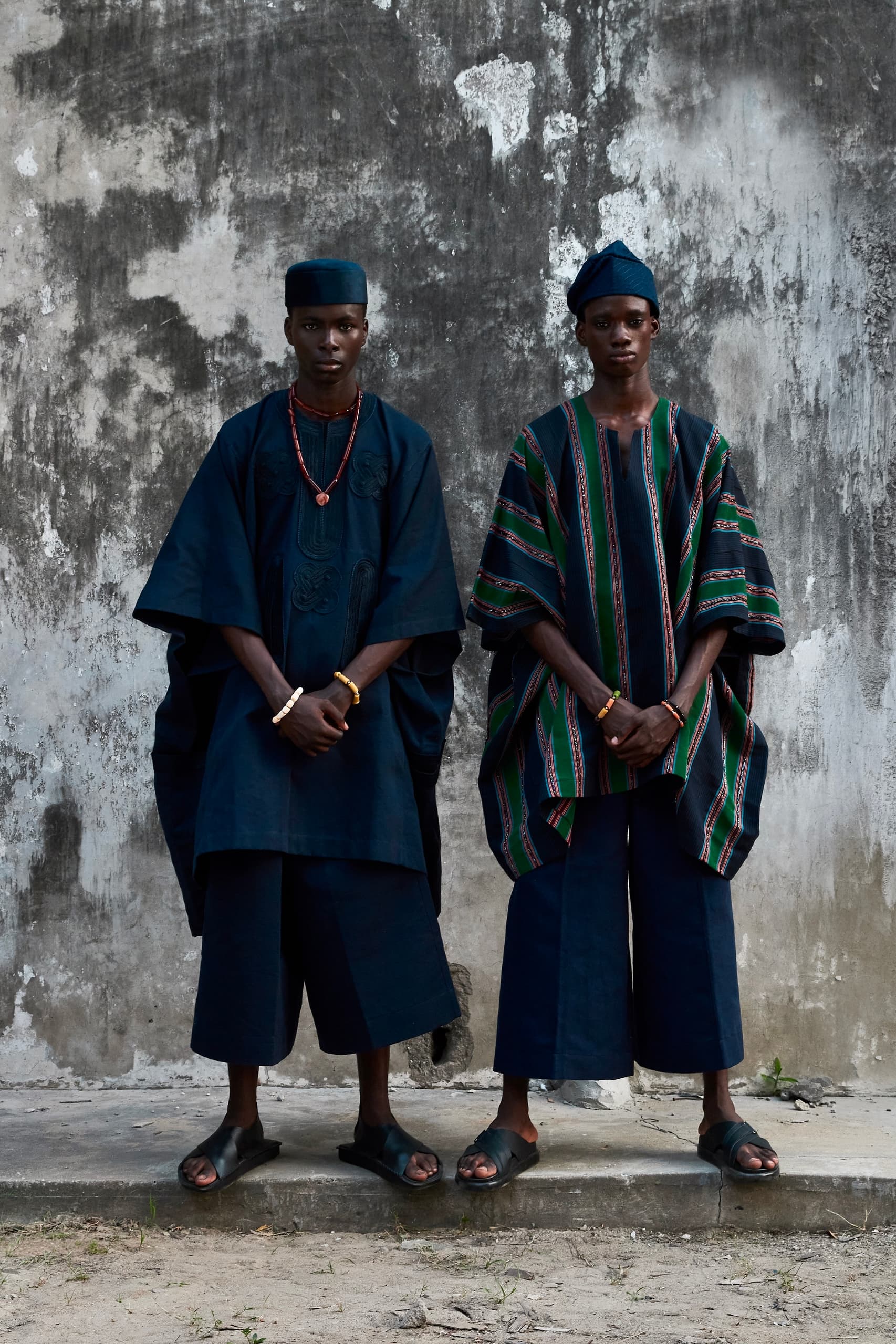 Two individuals in traditional African attire standing against a textured wall.
