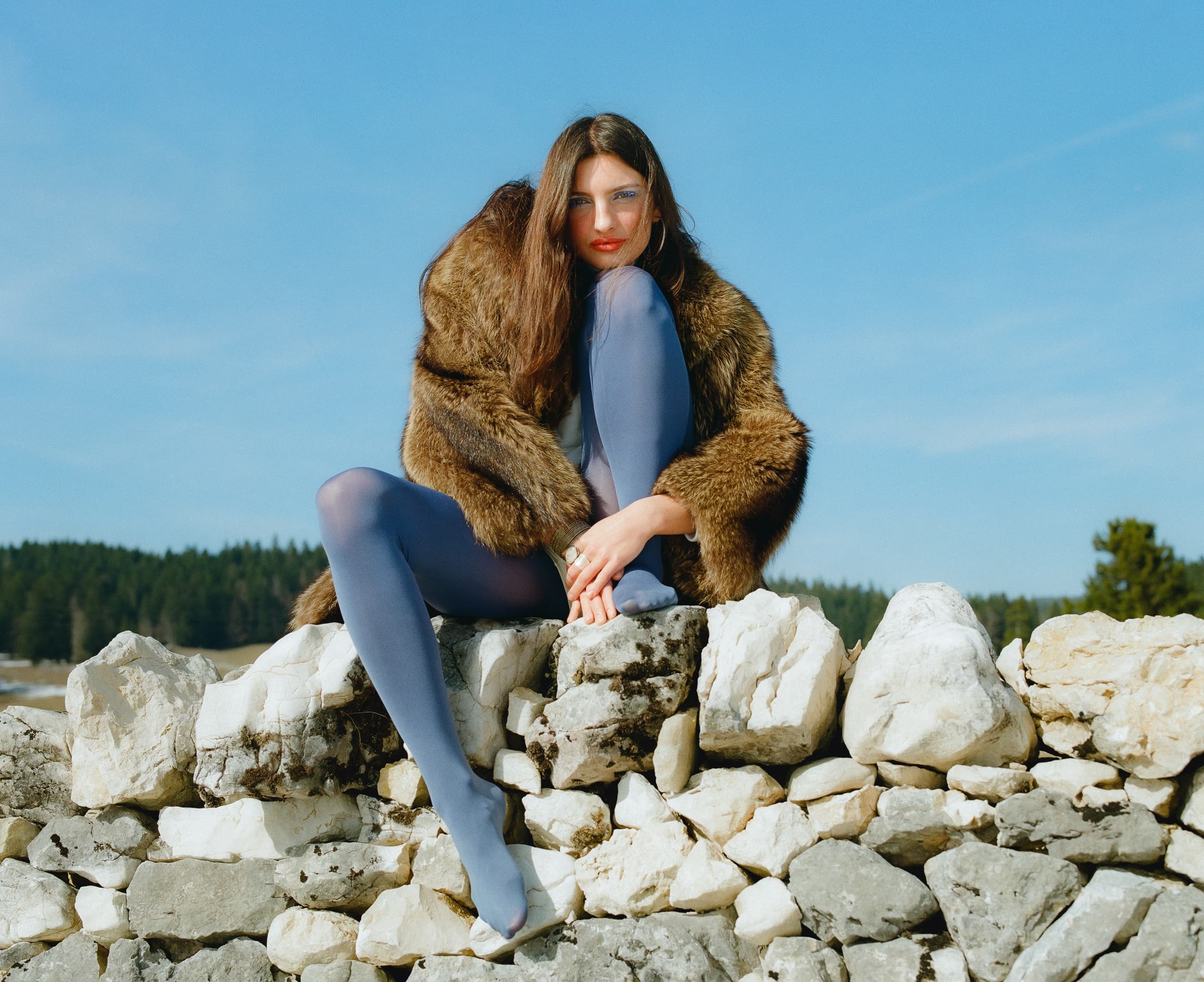 Woman in fur coat and blue tights sitting on a stone wall outdoors.