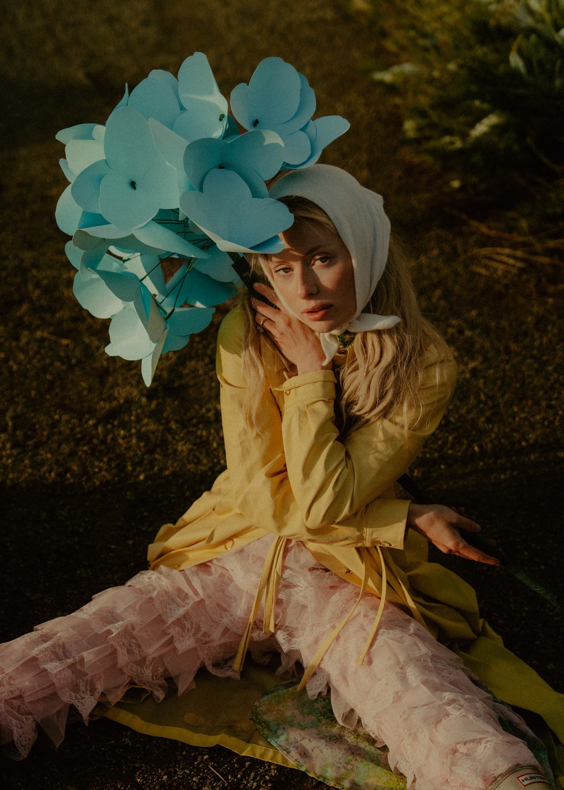 Woman in yellow coat with blue paper flowers and white headscarf in garden setting.