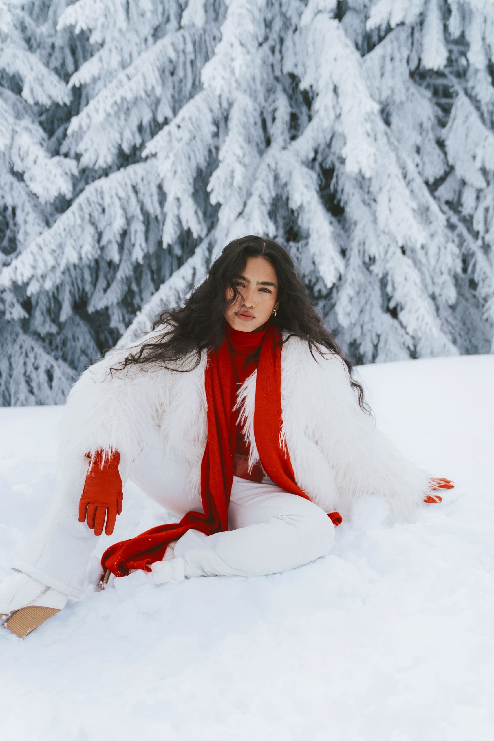 Woman in winter fashion sitting in snowy forest with red scarf and white coat.