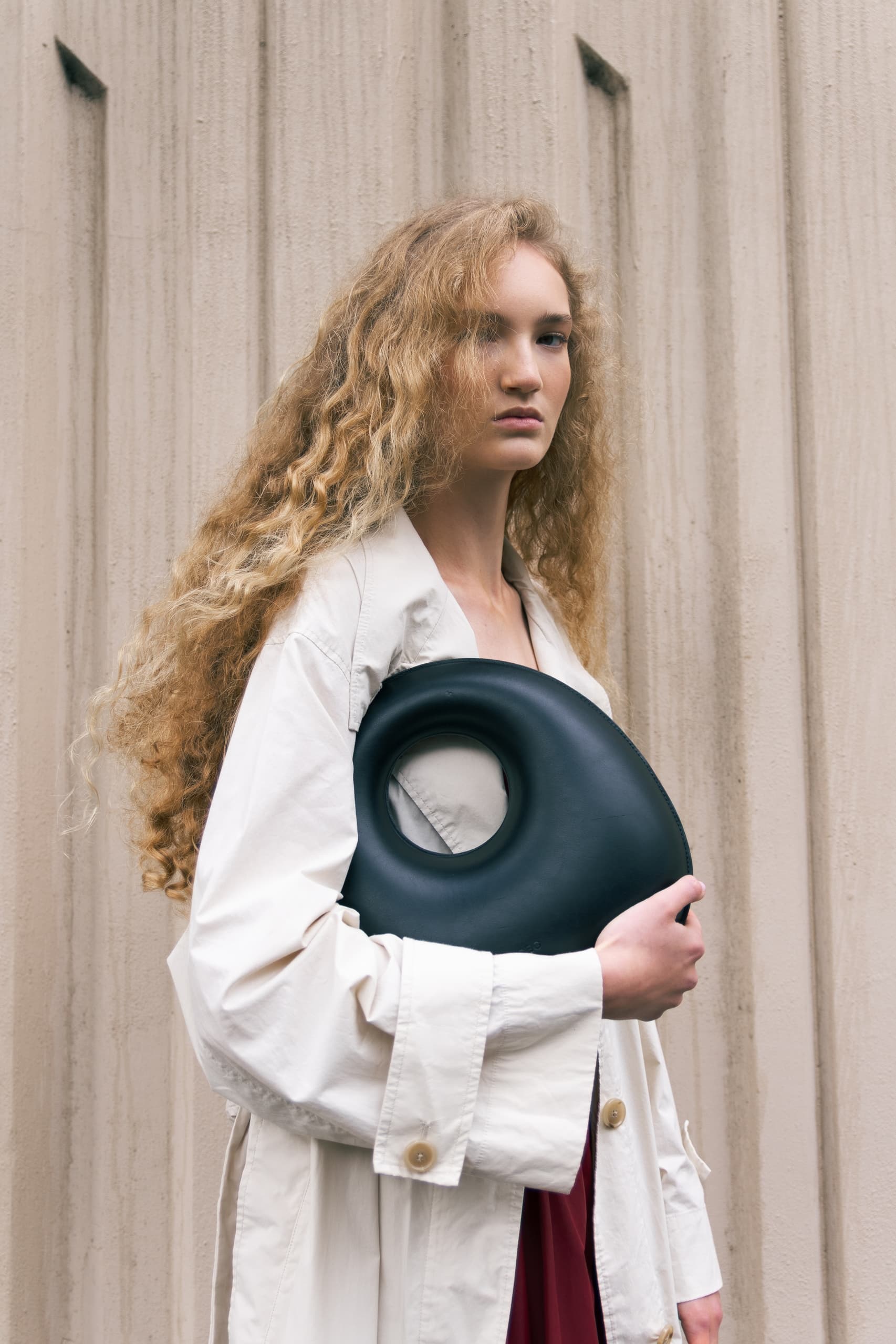 Trendy woman in beige coat holding black circular bag against textured wall.
