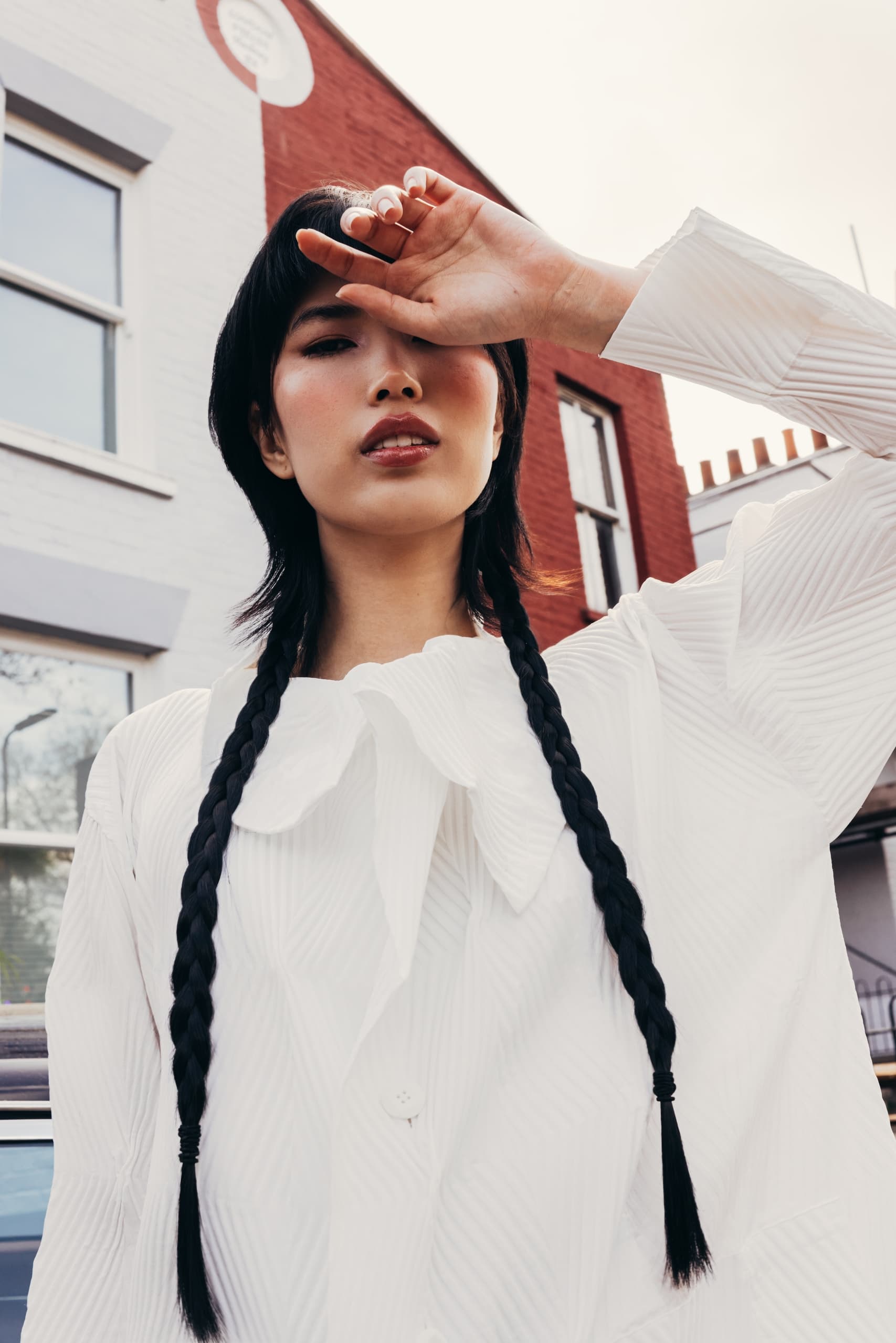 Woman in white shirt with long braids poses outside, shielding eyes from sun.