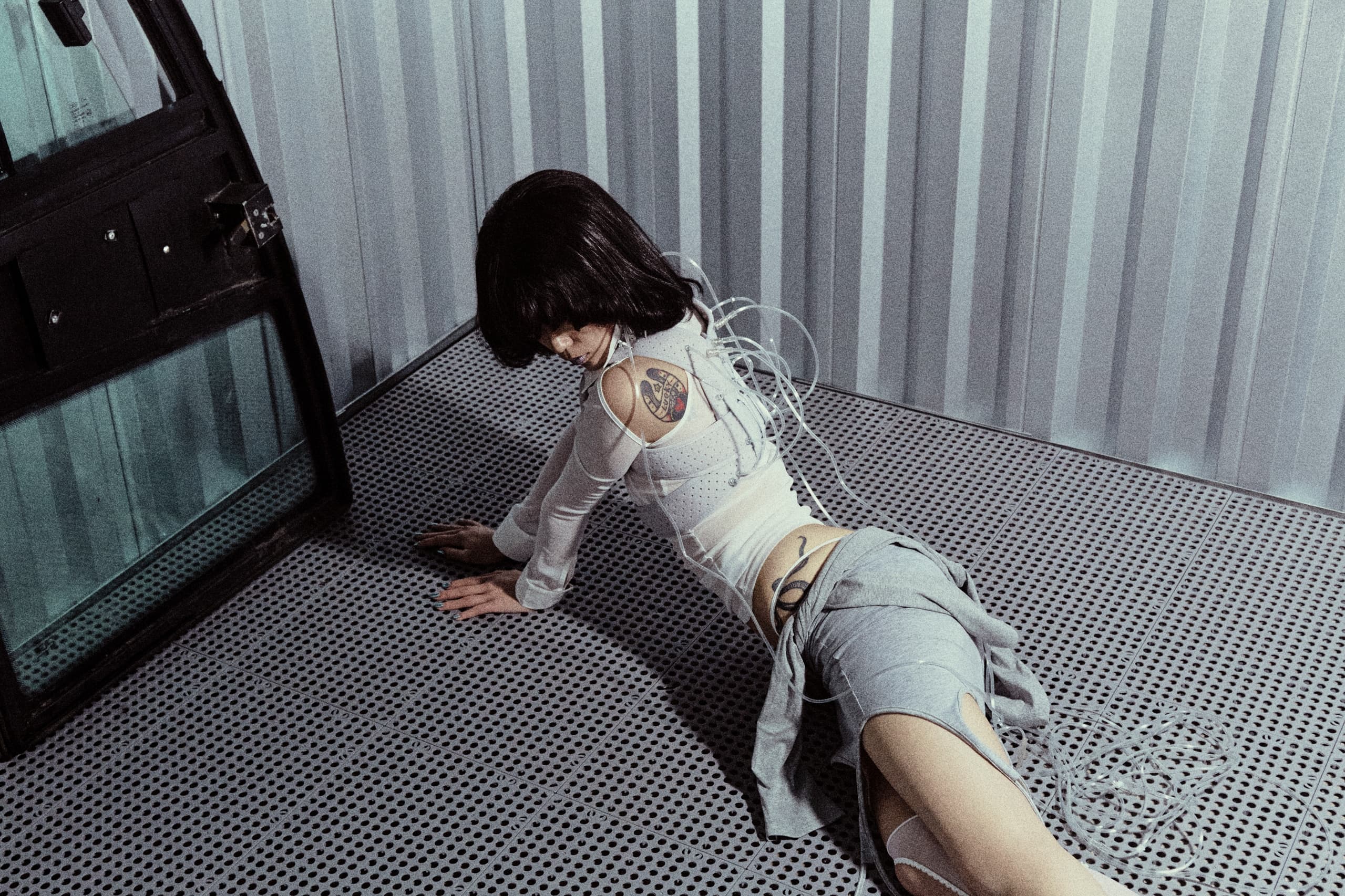 Futuristic scene: woman with tattoos and cables kneeling on metal floor.