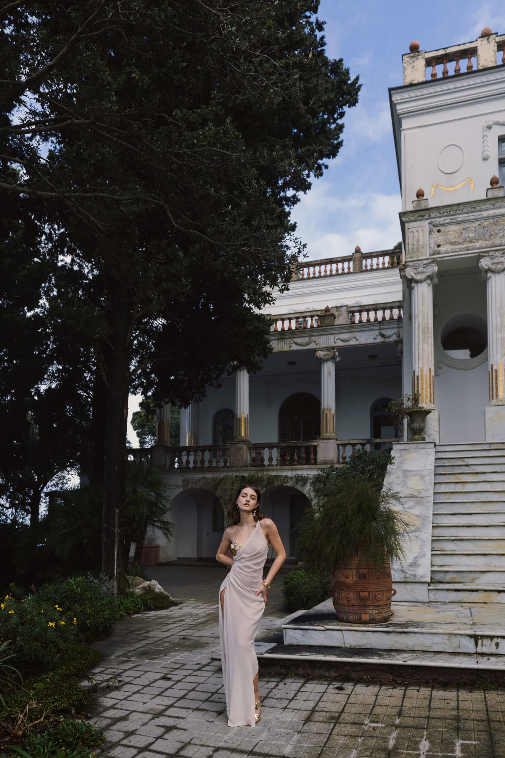 Elegant woman in evening gown by a grand estate under tall trees.