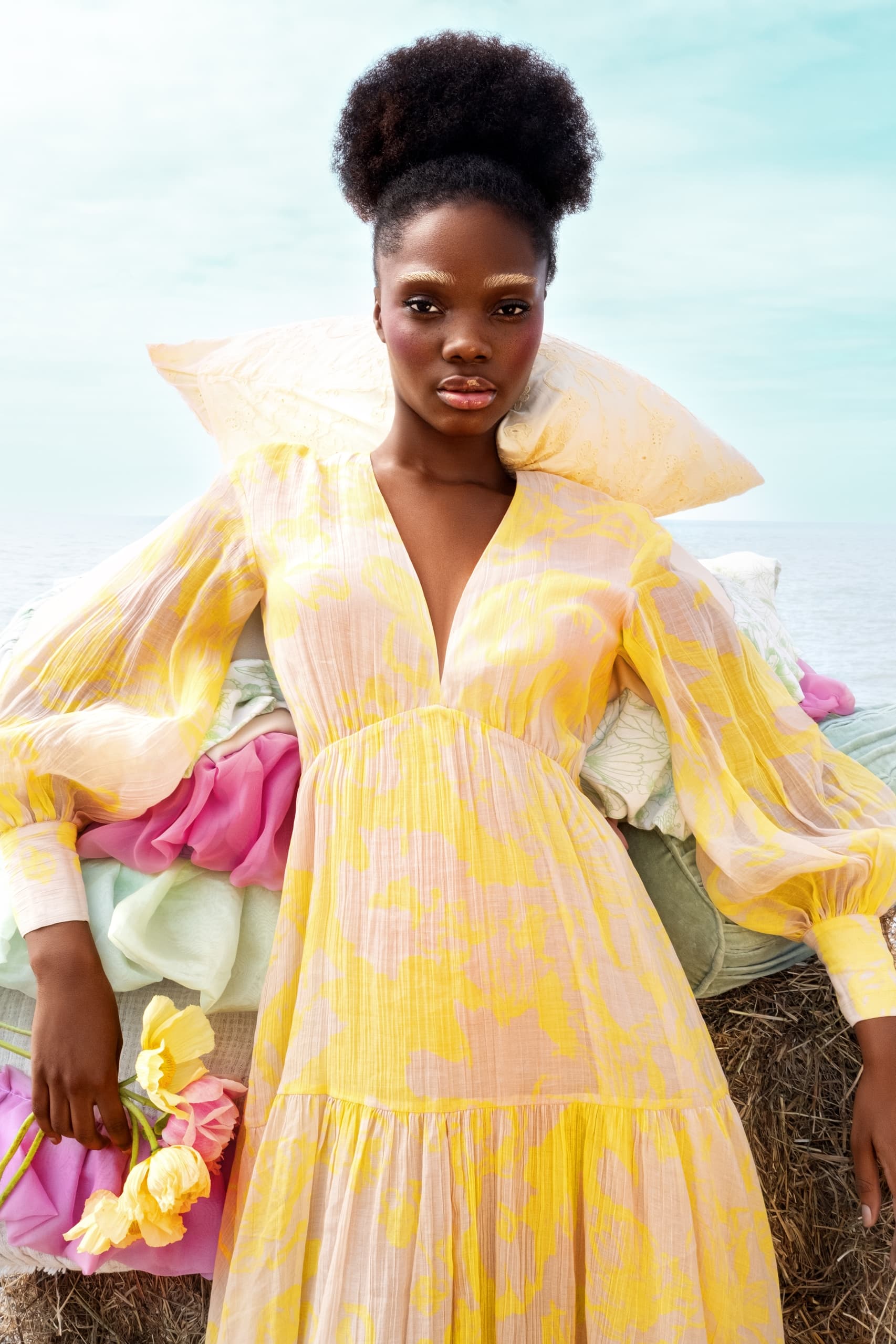 Woman in yellow floral dress holding flowers by the seaside.
