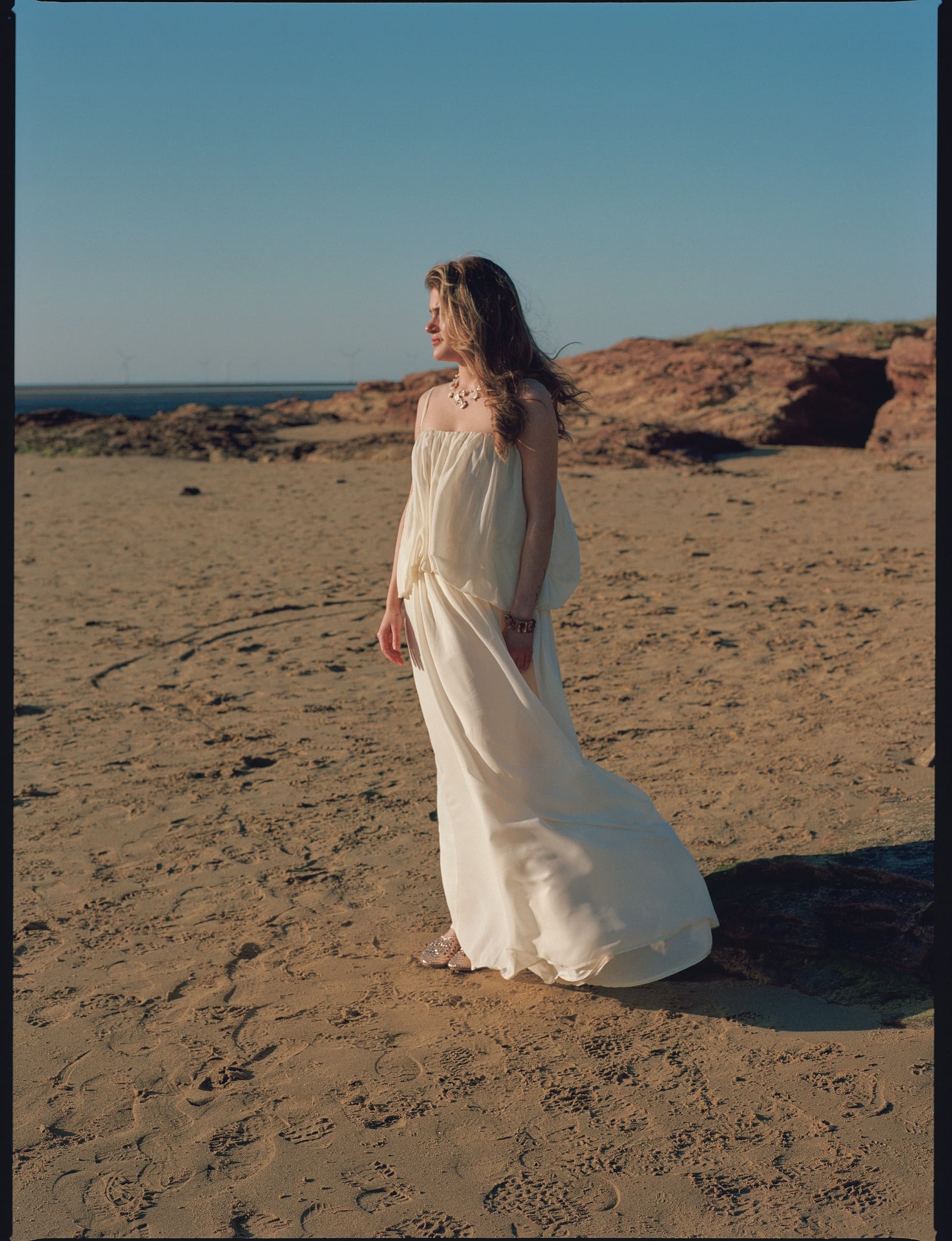 Woman in flowing dress stands on sandy beach under clear blue sky.
