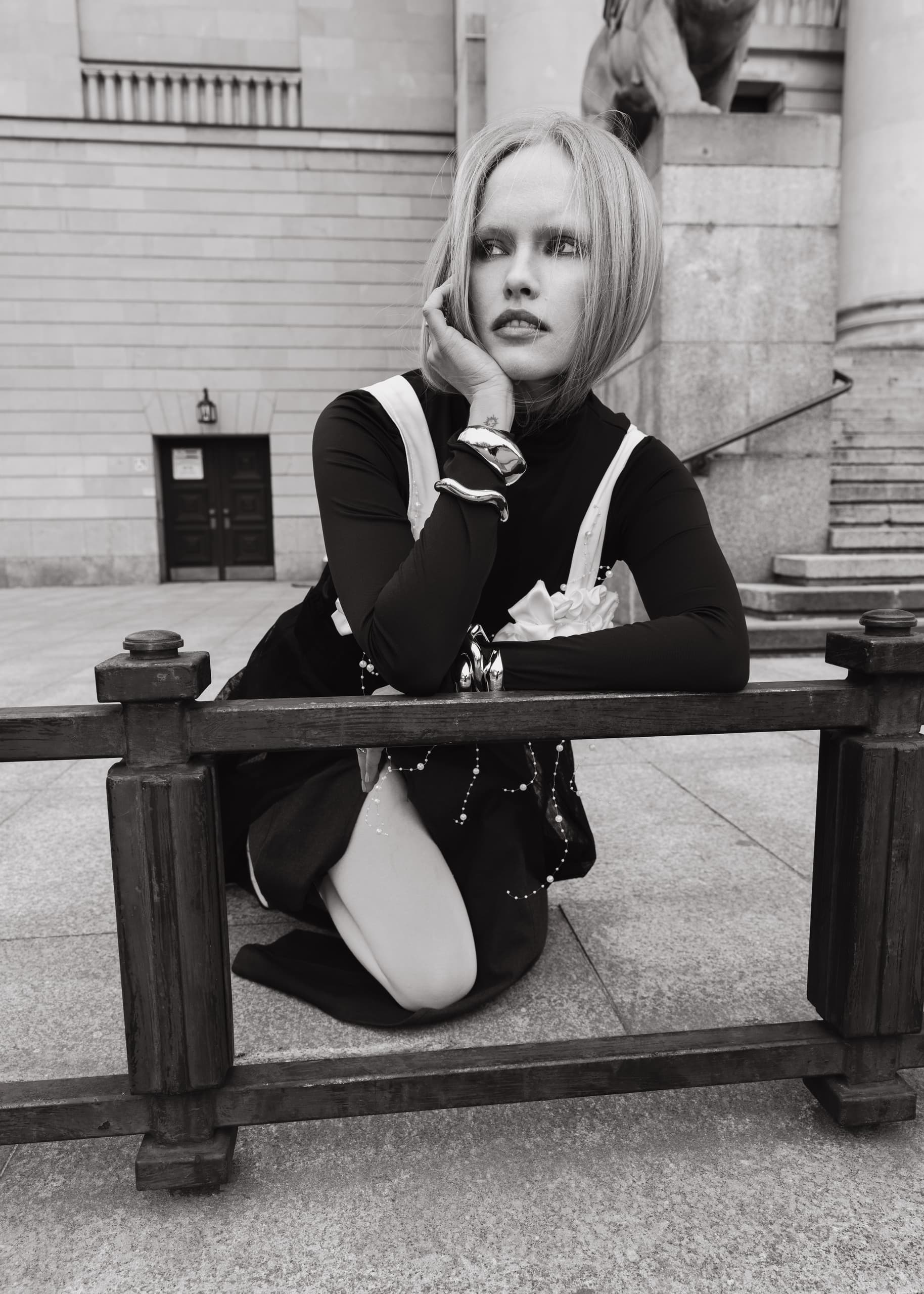 Fashion model in black and white attire poses artistically by a fence.