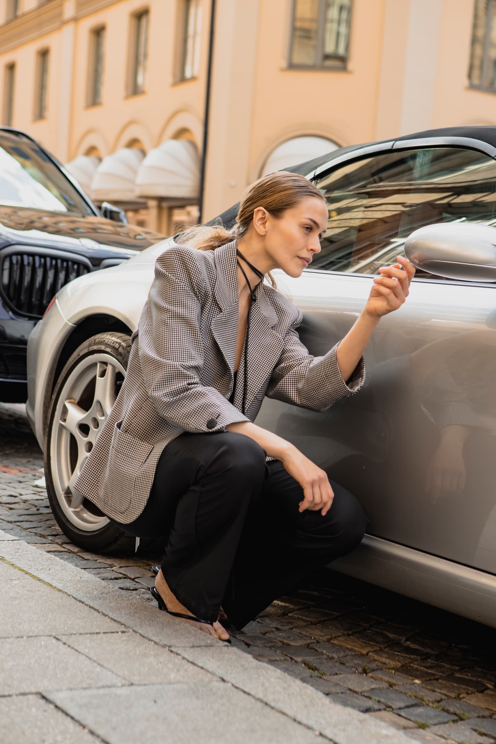 Woman in stylish blazer examines her reflection in a car's side mirror.