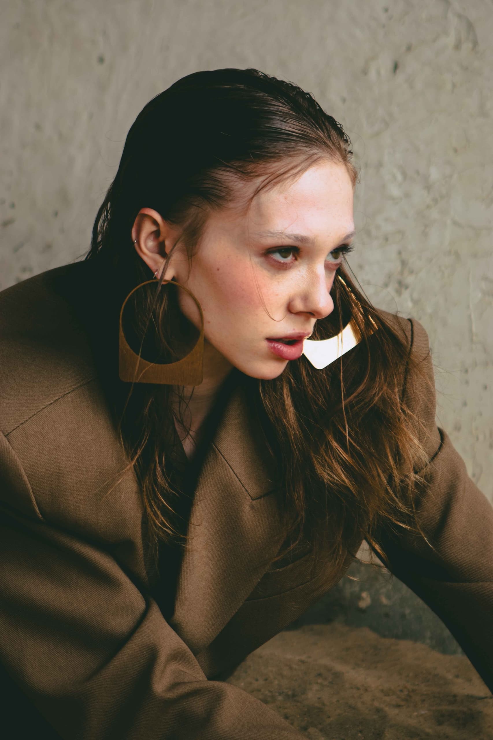 Woman in brown blazer with large geometric earrings, posing against textured wall.