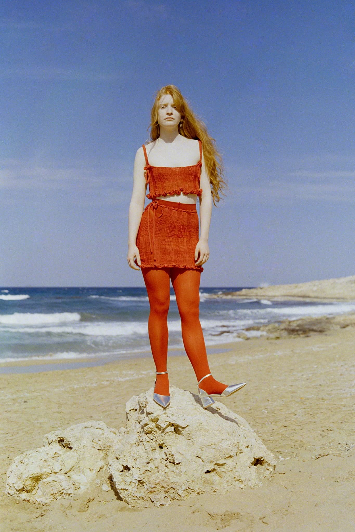 Woman in red dress standing on beach rock under a clear blue sky. Fashion photo.