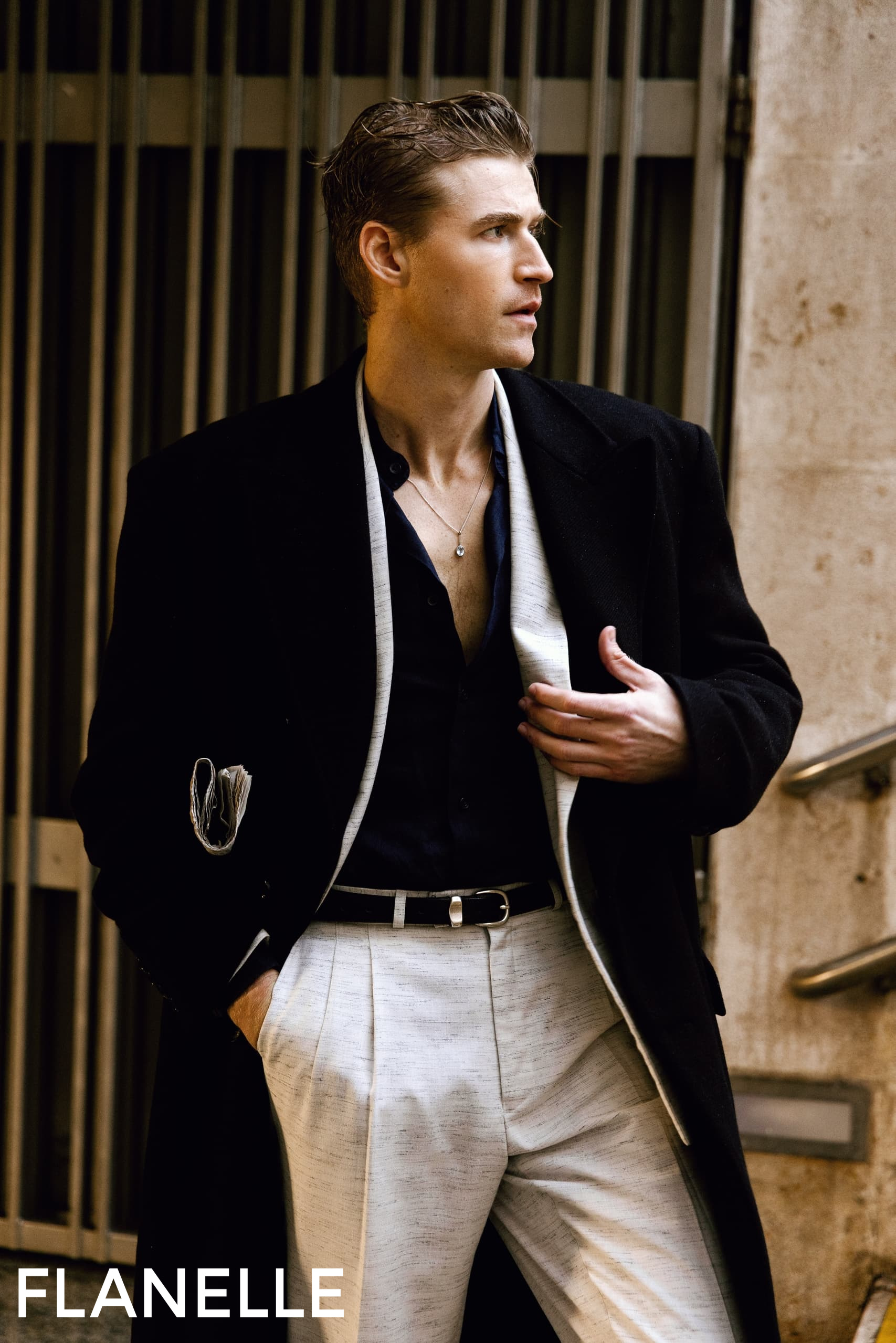 Man in stylish black coat and white pants, urban backdrop, Flanelle fashion brand.