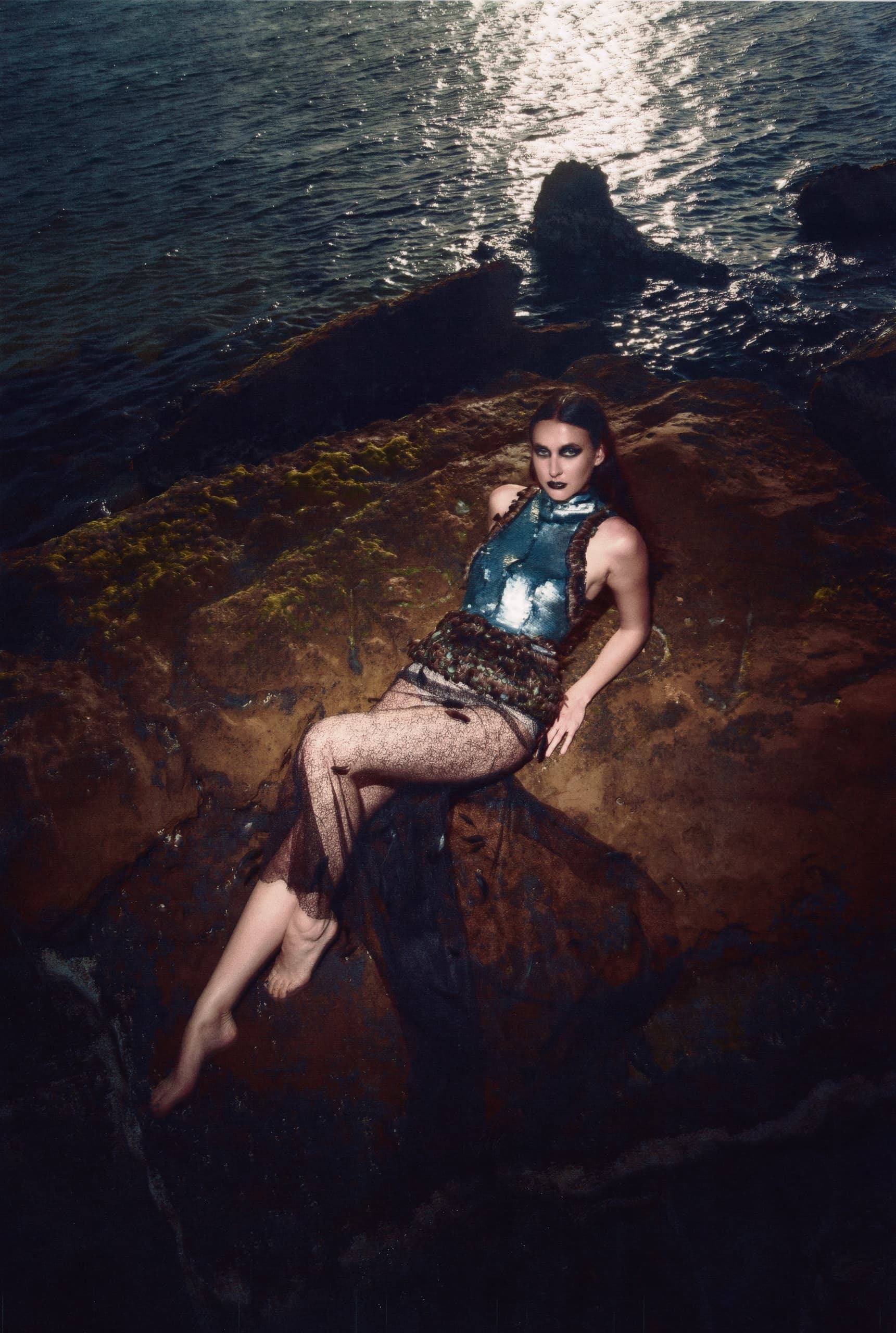 Model in artistic pose on rocky shore with shimmering water in background.