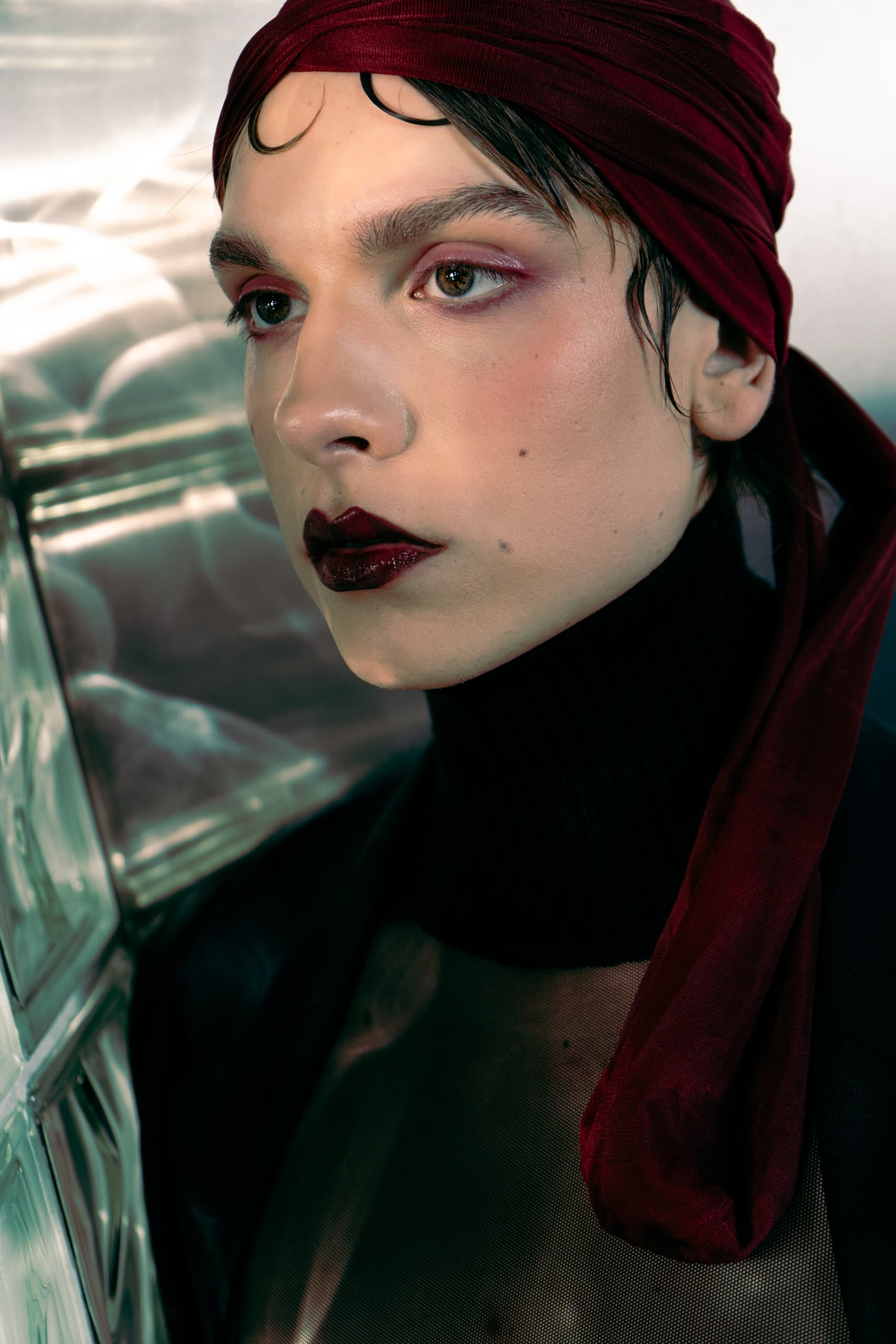 Person with bold makeup and red headscarf, standing near glass tiles.