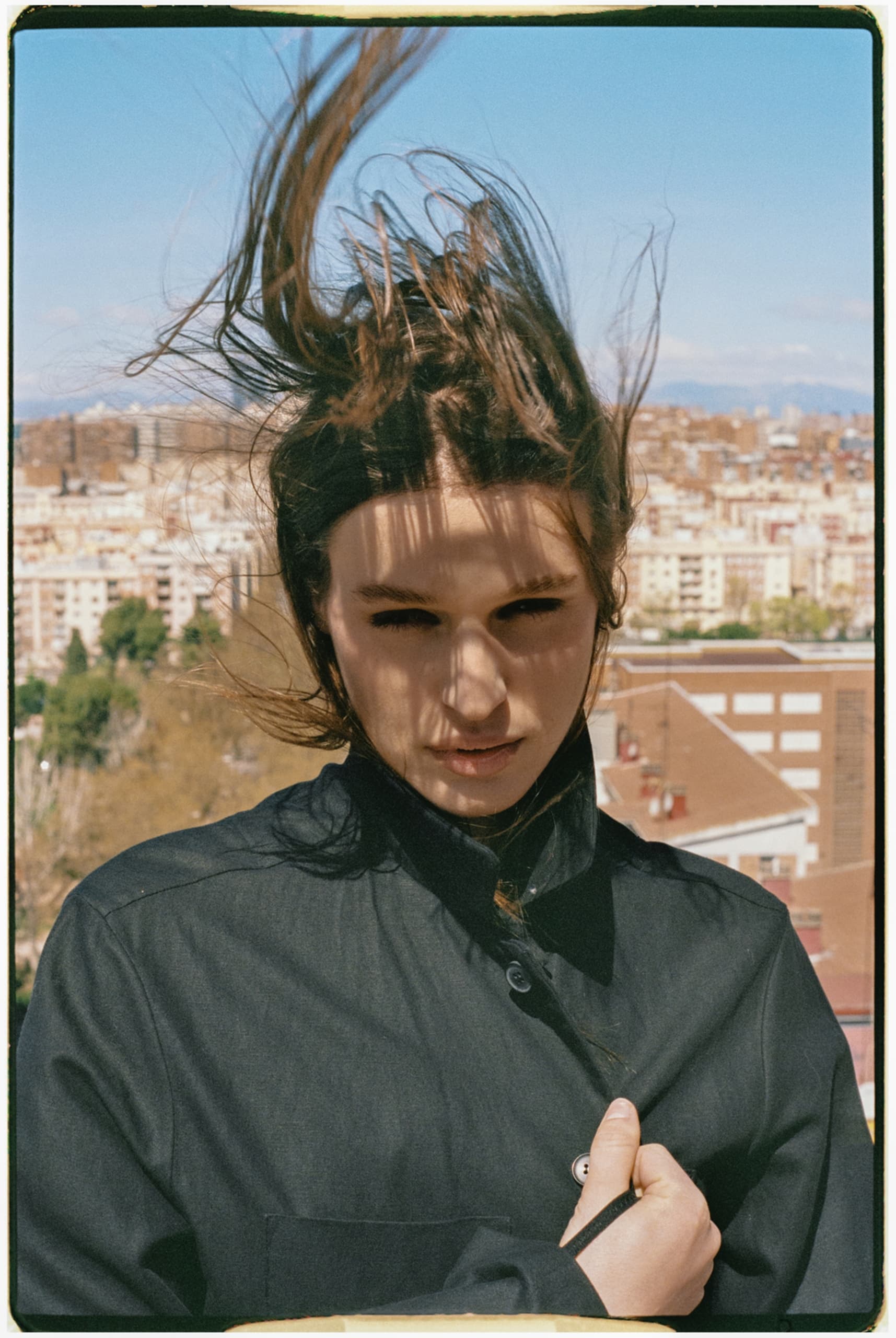 Person with windswept hair in urban setting, wearing a black jacket.