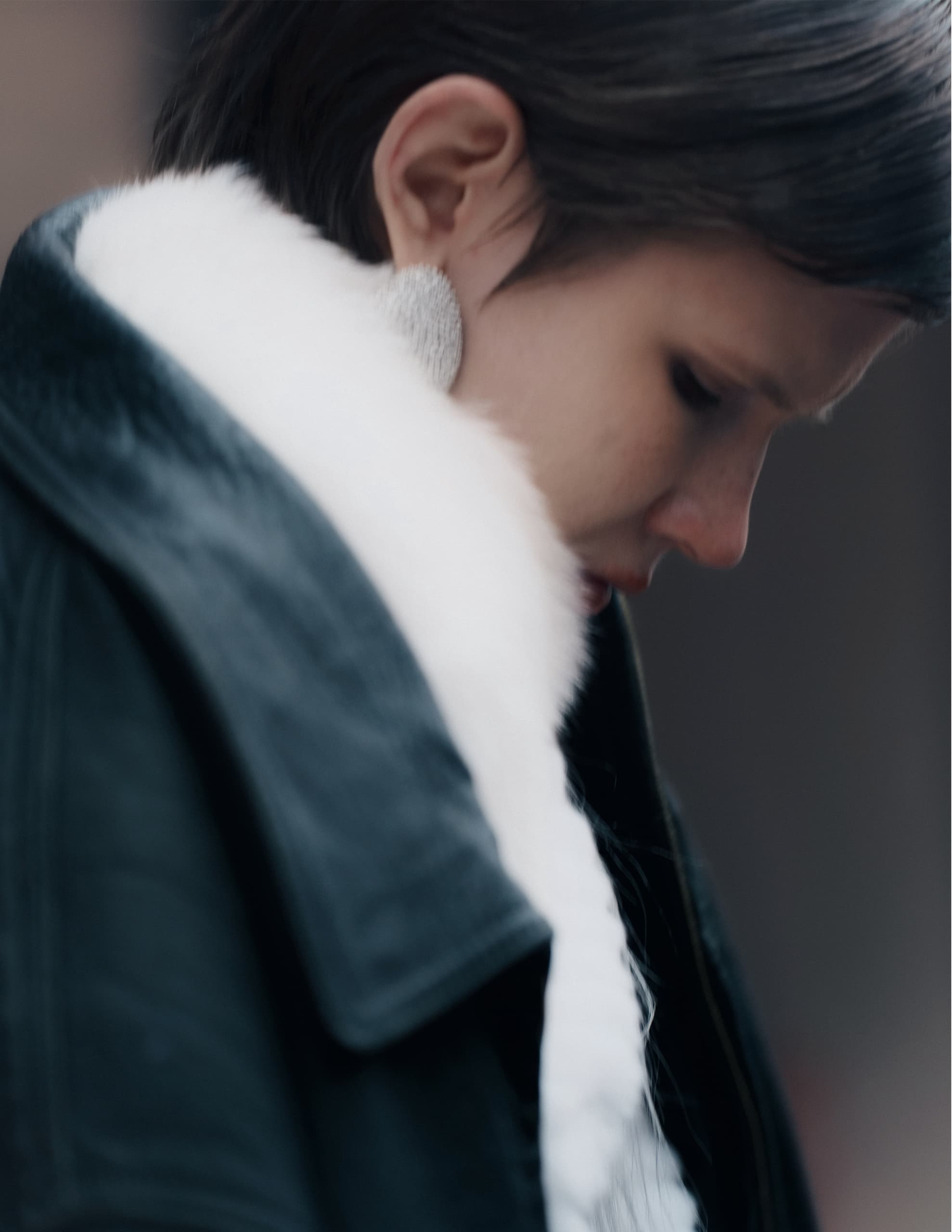 Stylish person in fur coat and silver earrings, looking down thoughtfully.