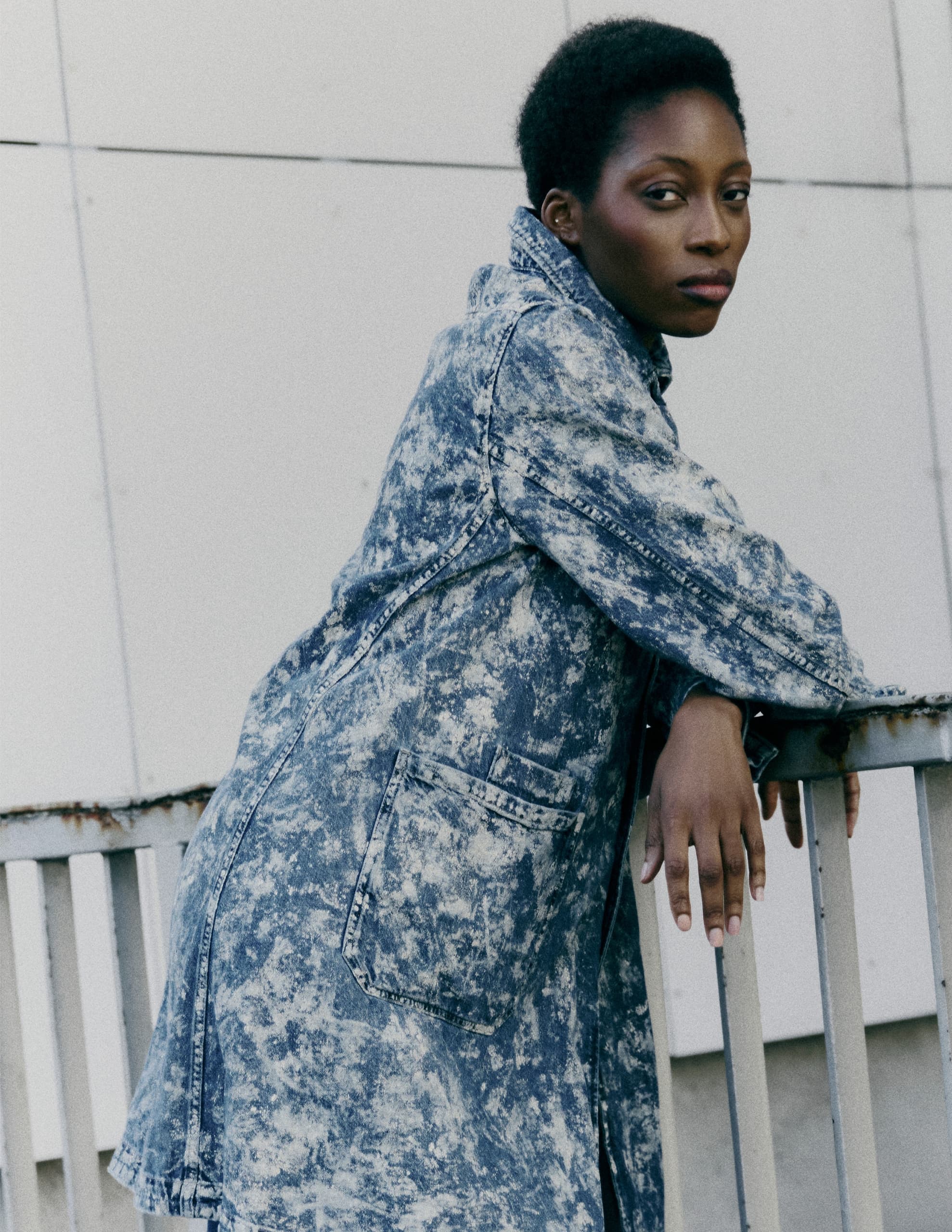 Fashionable person in blue acid-wash denim coat, leaning against a metal railing.