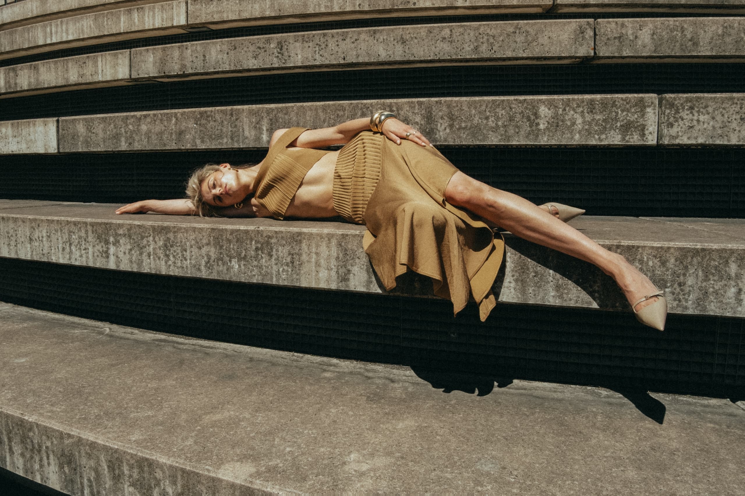 Fashionable woman in beige outfit reclining on concrete steps outdoors.