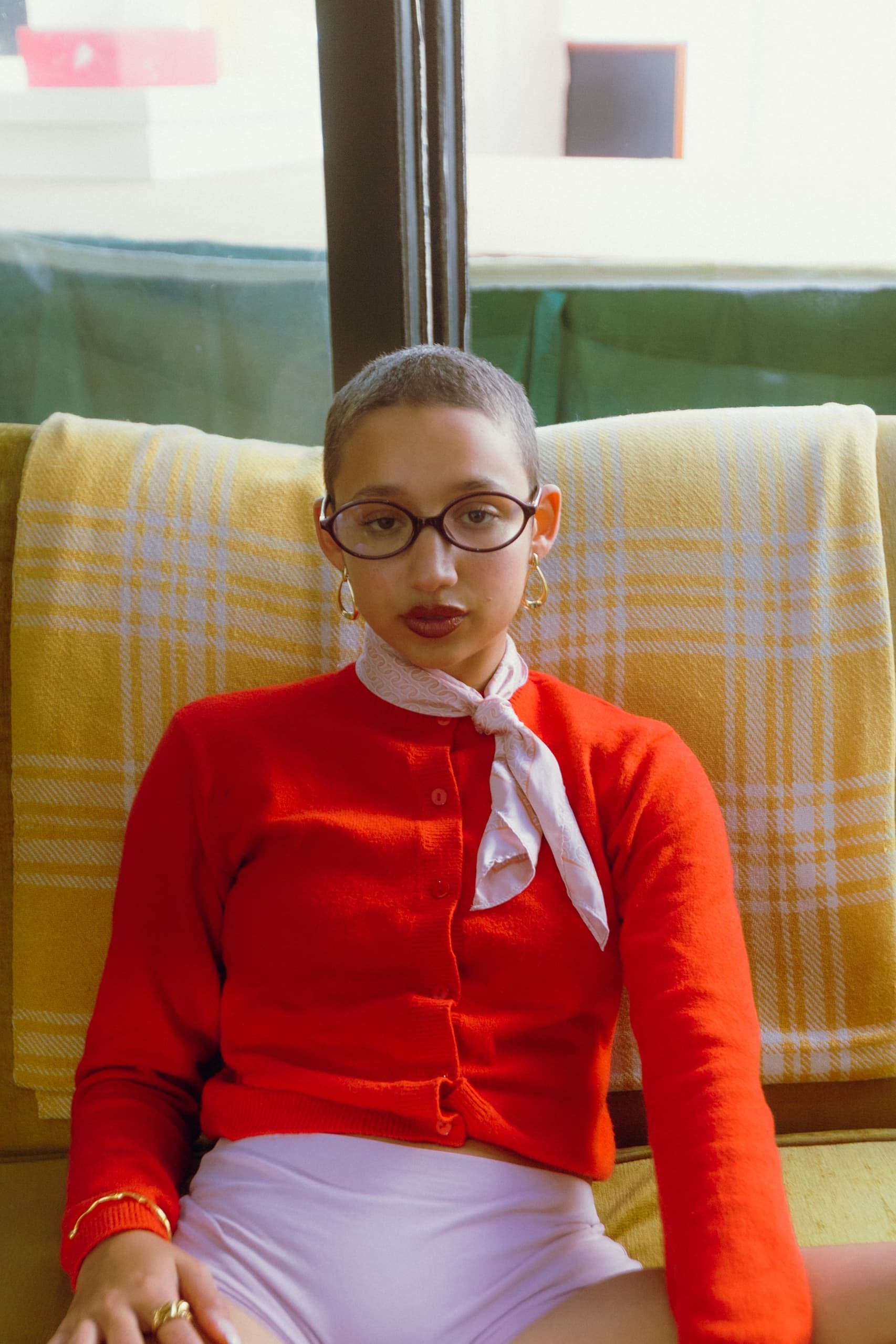Person in red sweater sitting indoors, stylish with glasses and scarf.