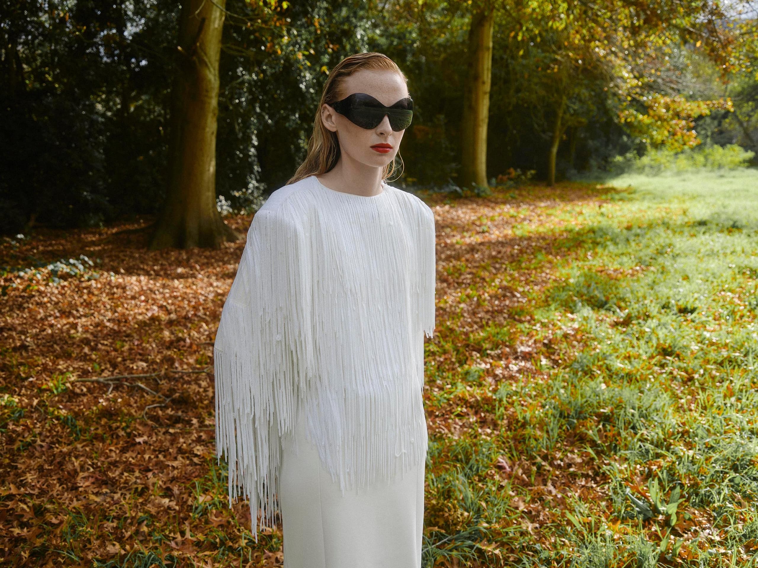 Woman in fringe white dress and sunglasses stands in sunlit forest clearing.