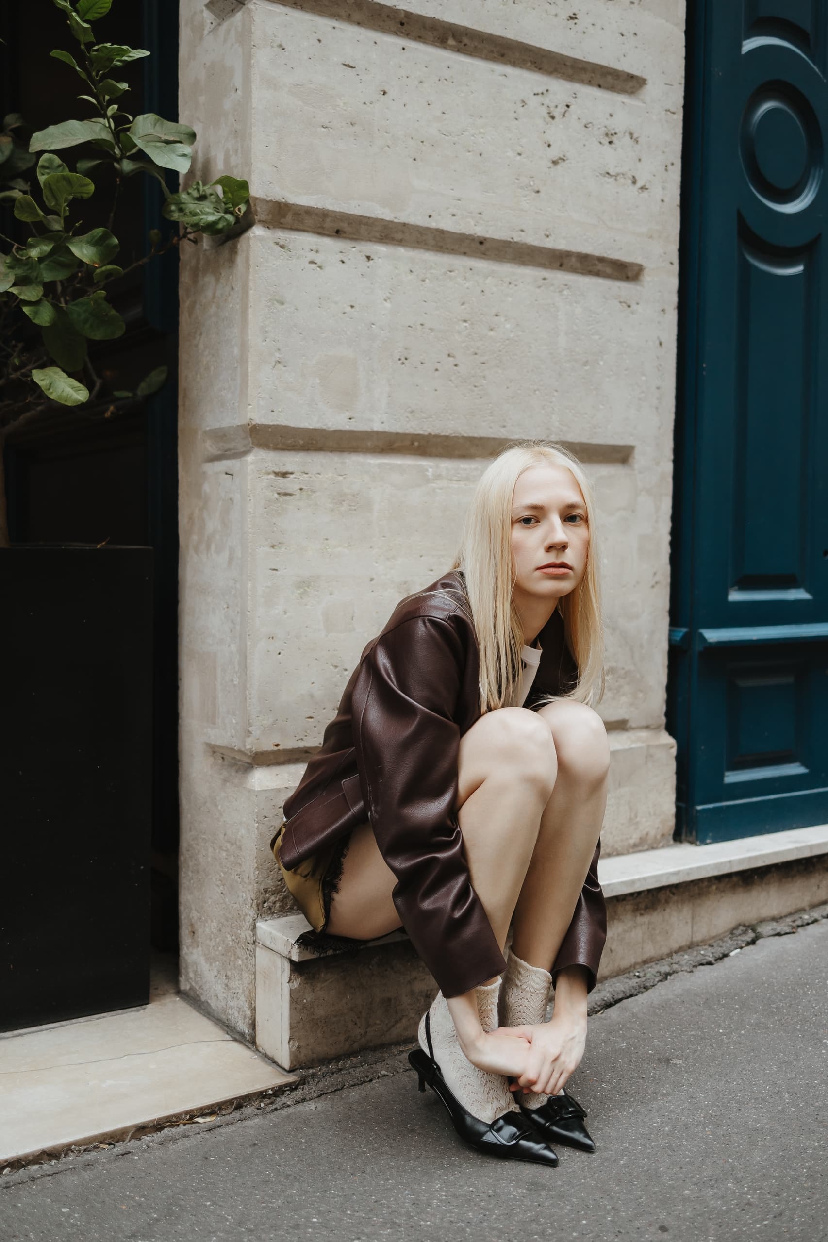 Person in brown jacket sitting on steps near a door in a city street setting.