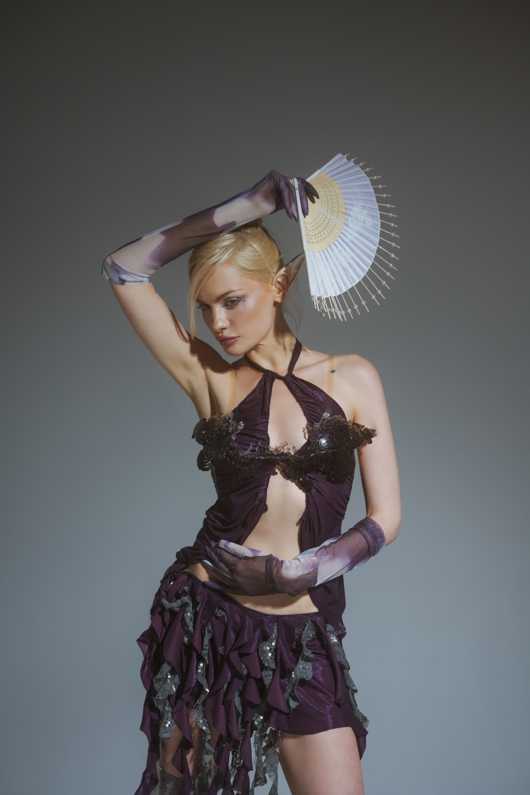 Elegant woman in purple attire holding a decorative fan against a gray backdrop.