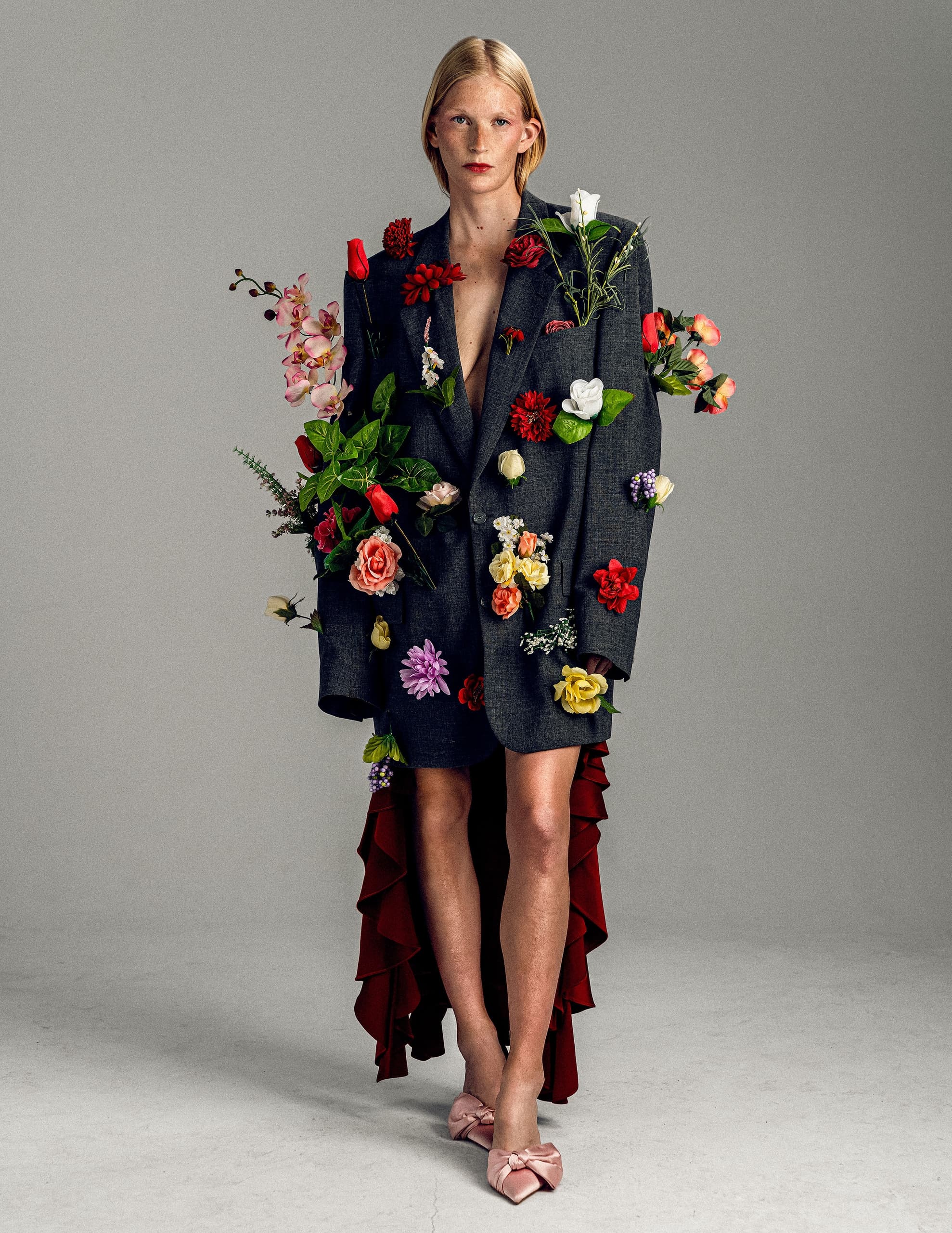 Model in floral-decorated outfit with dramatic red train and pink heels. Fashion-forward look.