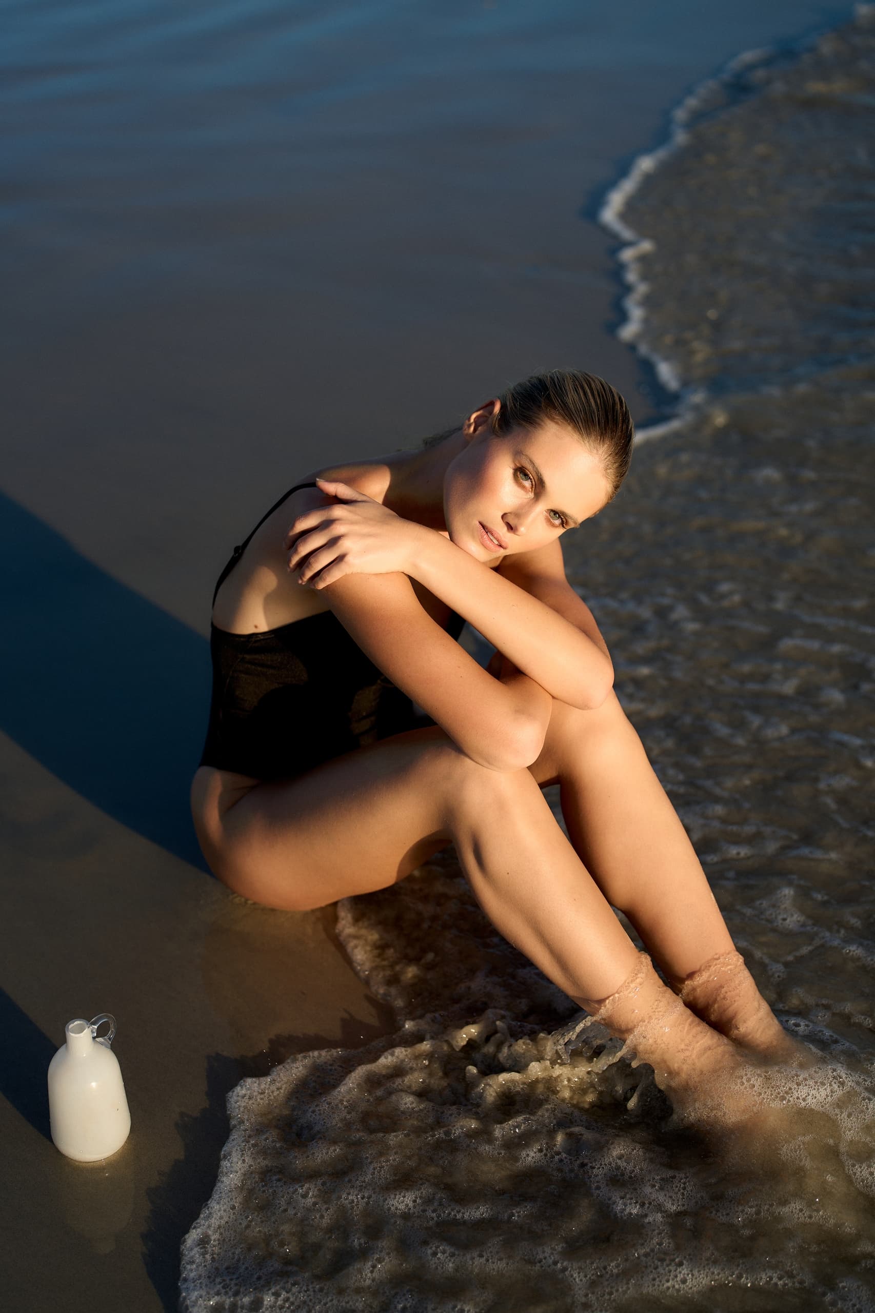 Woman in black swimsuit sitting on seashore with waves and a white jug nearby.