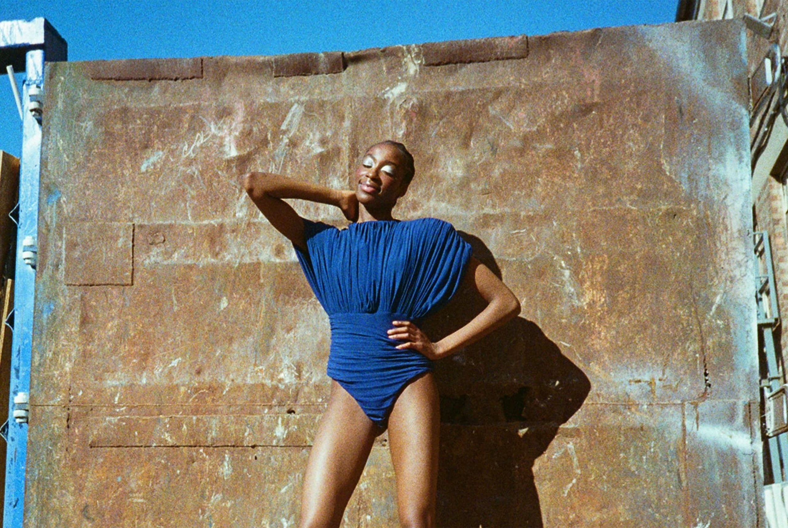 Person poses in a blue outfit against rustic metal background in sunlight.