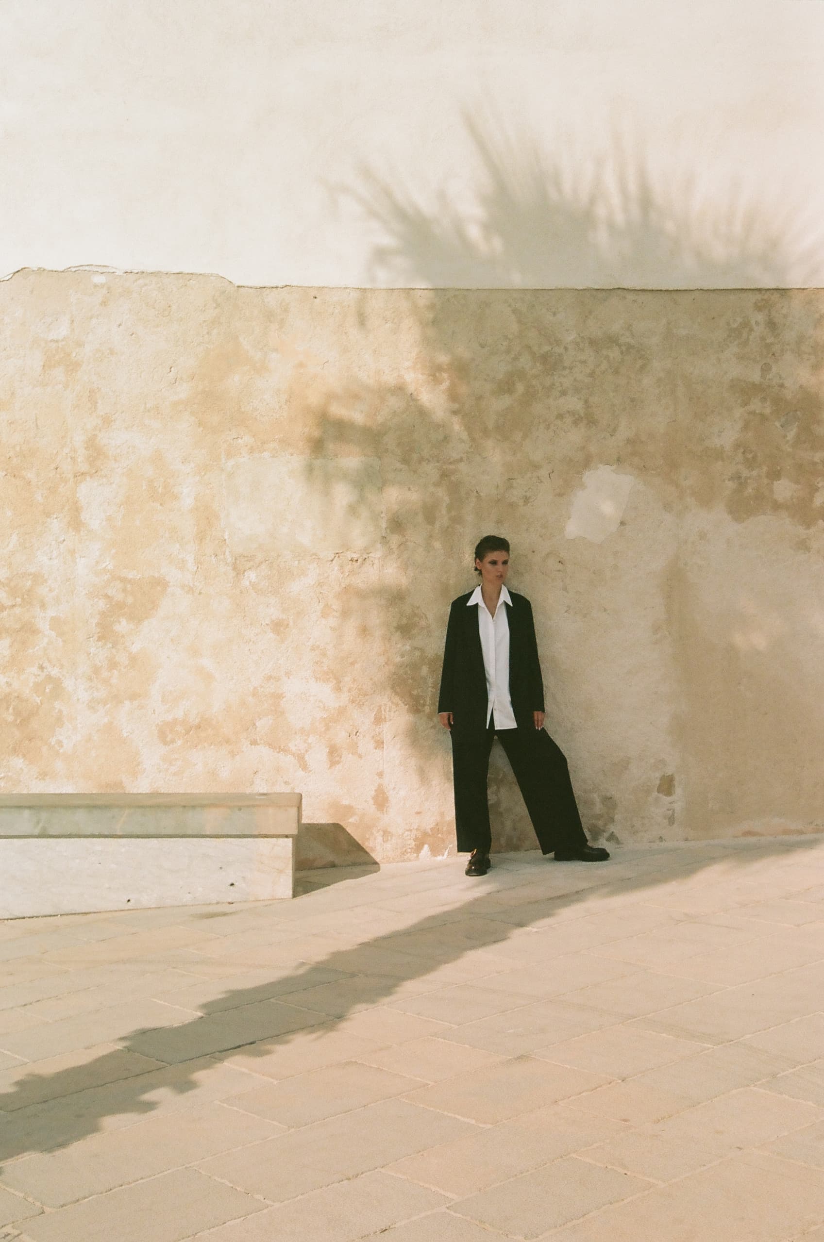 Person in black suit under palm shadow against textured wall.