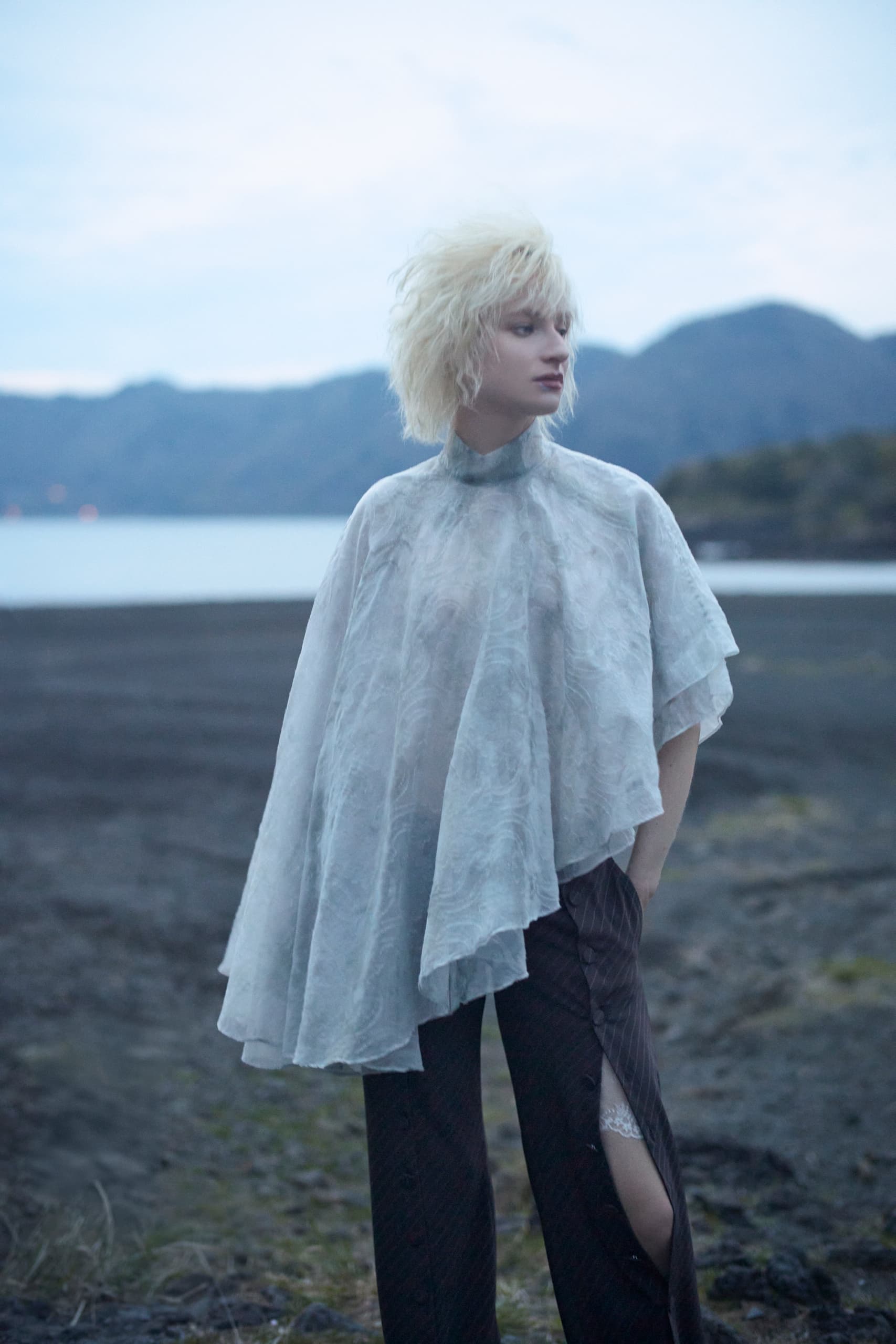 Person in sheer outfit stands by serene lake, mountains in the background.