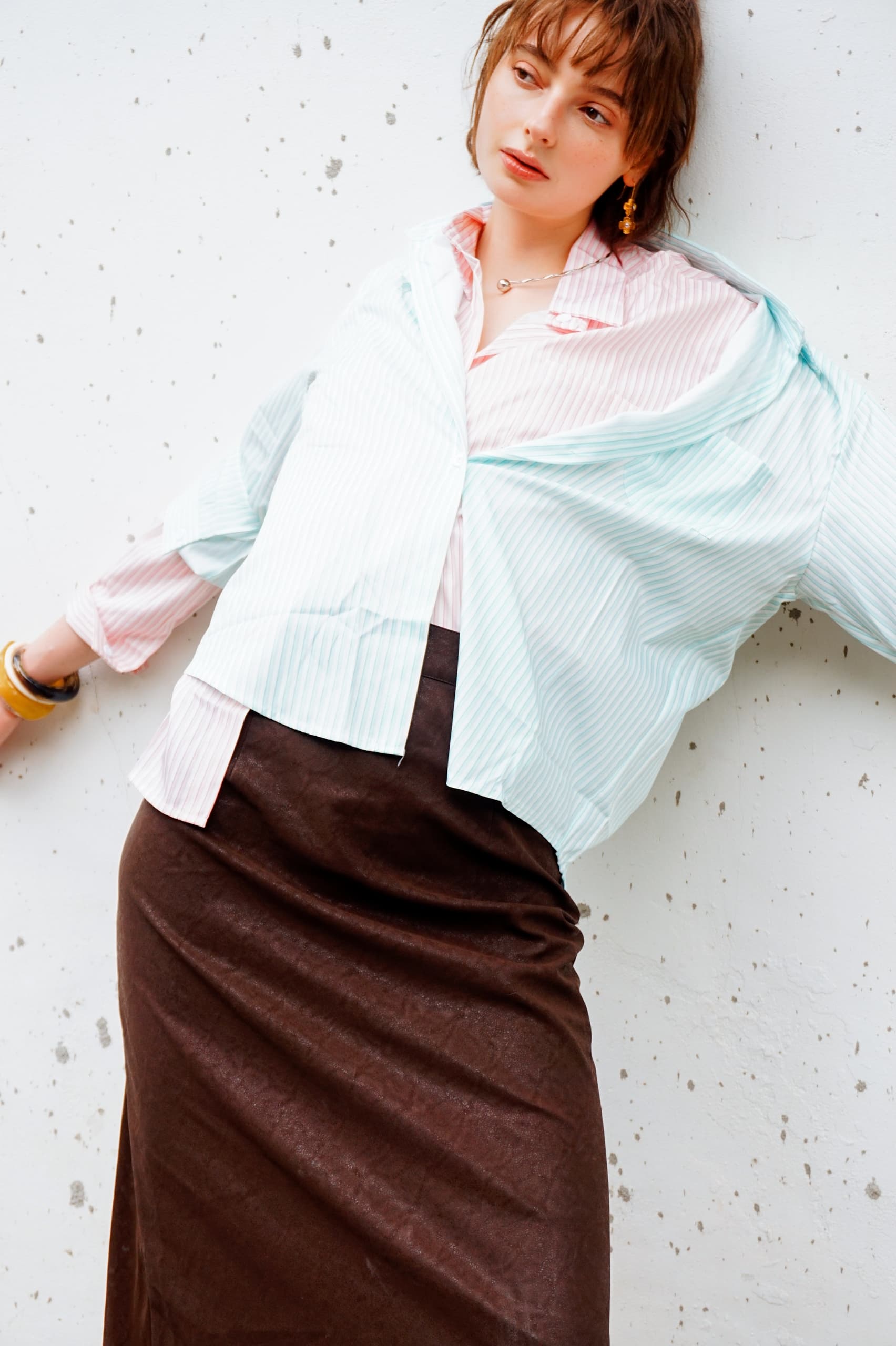 Fashionable woman in striped blouse and brown skirt leaning against a white wall.